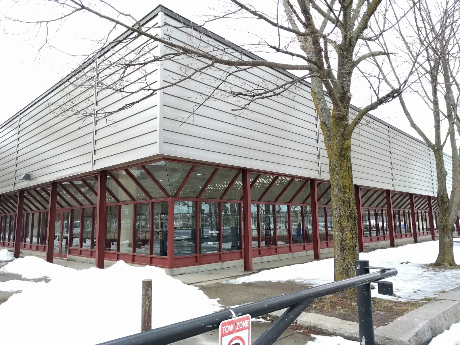 Corner view of the rink building's exterior in winter, with cream metal siding over a red-framed glass entry wall, snow piled along the sidewalk and a "Tow Zone" sign in front. — DCR Steriti Memorial Rink