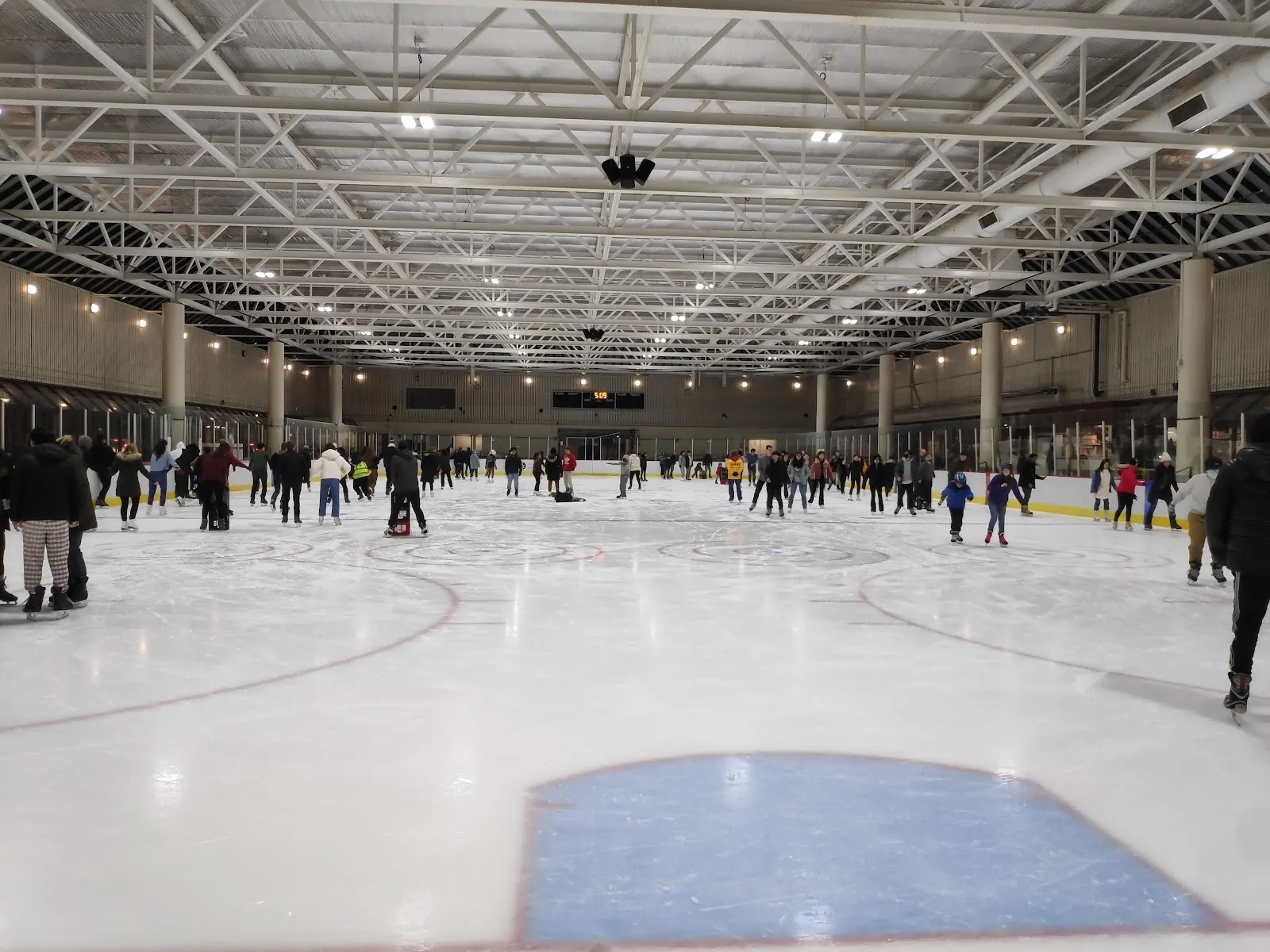 A public session on a wide covered sheet, skaters spread across the ice under a long steel roof with tall windows along the far side. — DCR Steriti Memorial Rink