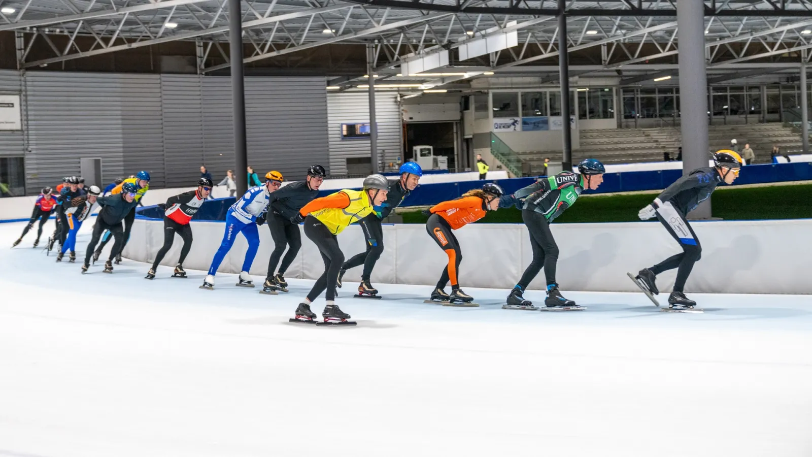 A group of speed skaters training in a pack along the straight of the De Meent ice track under the hall's steel roof. — IJsstadion De Meent