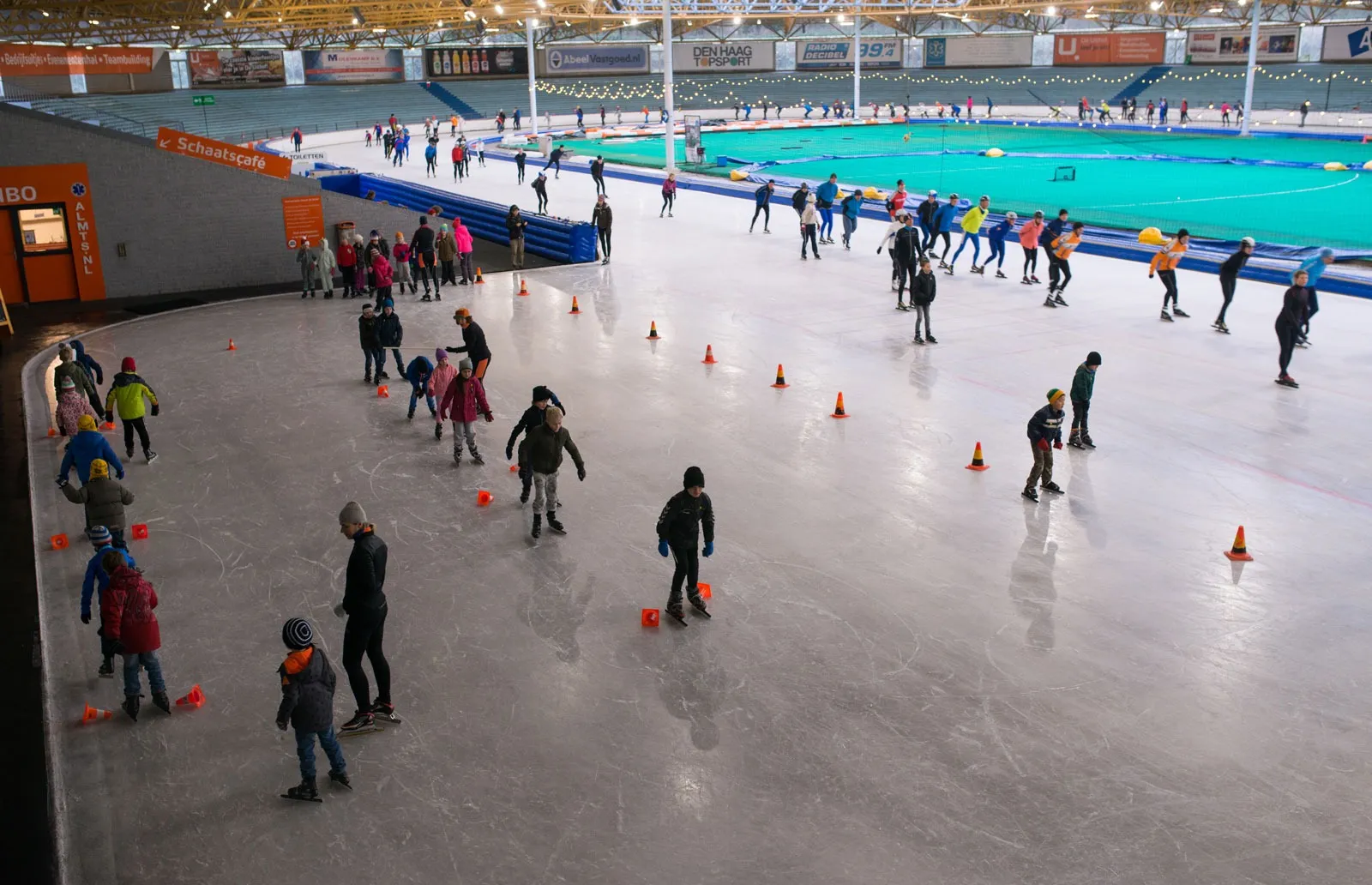 A public session at De Uithof, skaters spread over the wide ice with cones laid out and the green infield beyond. — De Uithof