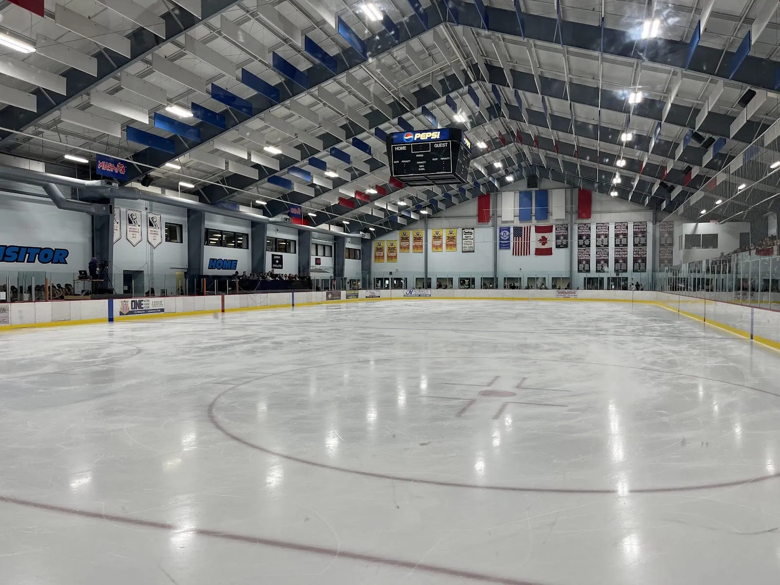 La pista principal de Dearborn desde el centro del hielo, vacía bajo los paneles rojos, blancos y azules del techo, con el marcador de Pepsi y las mantas de campeonatos — Dearborn Ice Skating Center