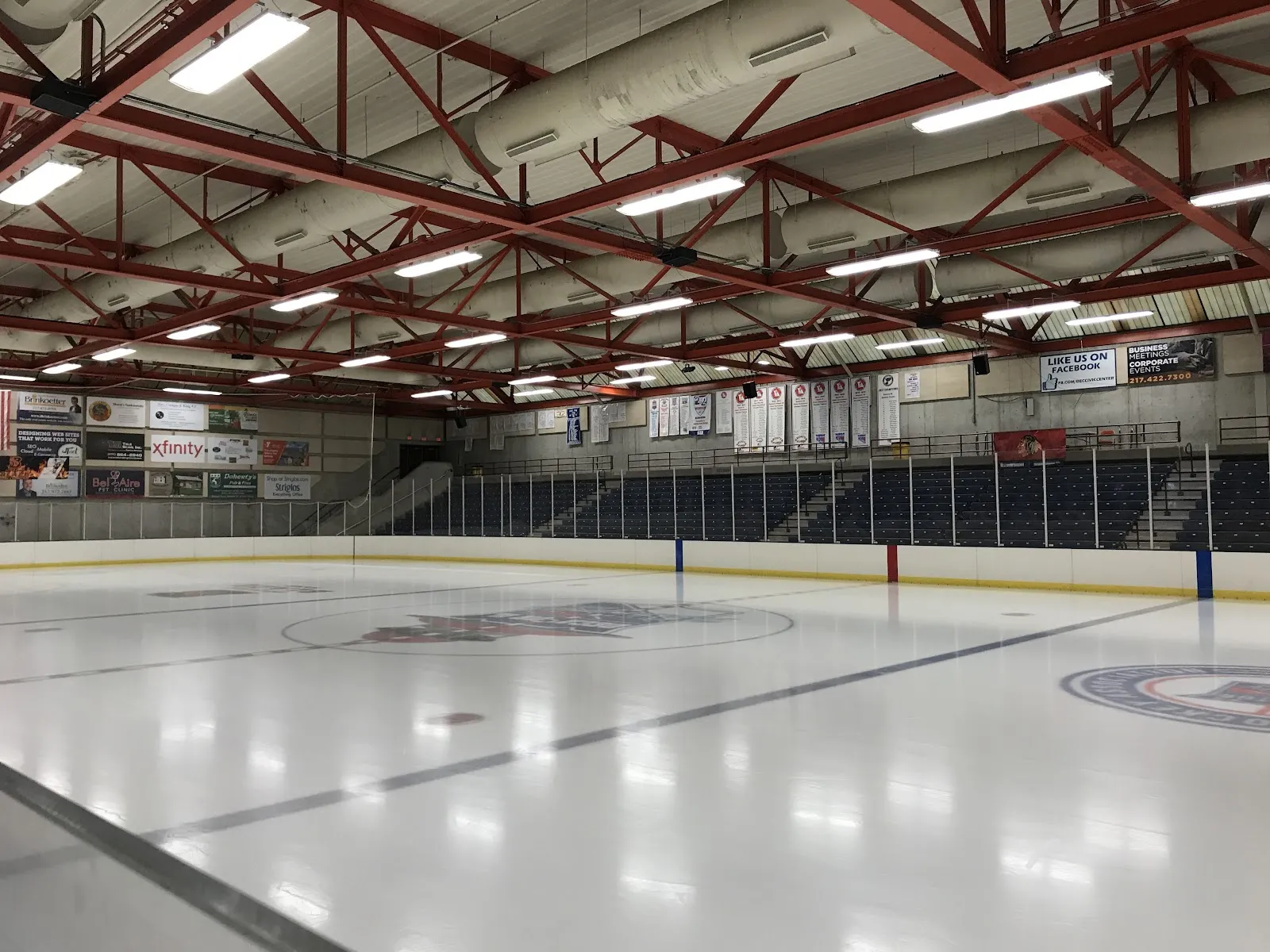 Empty full ice sheet at Decatur Civic Center, red steel roof trusses overhead, tiered navy seating and championship banners along the far side, center-ice and blue-line logos visible on the ice. — Decatur Civic Center