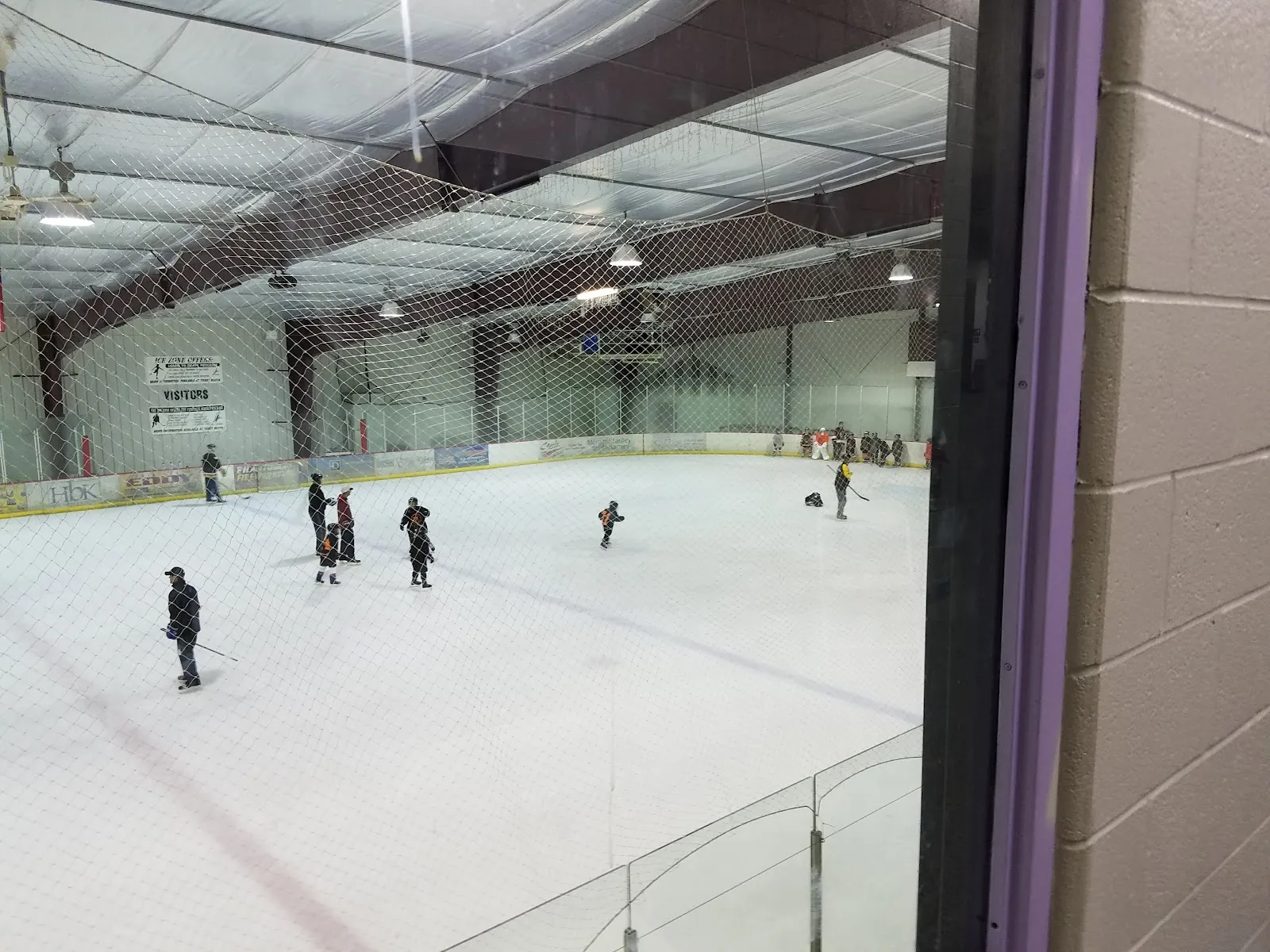 La pista de hielo vista a través de una red protectora y un marco de ventana desde un lado, con unos pocos patinadores repartidos en los extremos cercano y lejano, con bandas y una pared de acero clara al fondo. — Deep Freeze Ice Arena