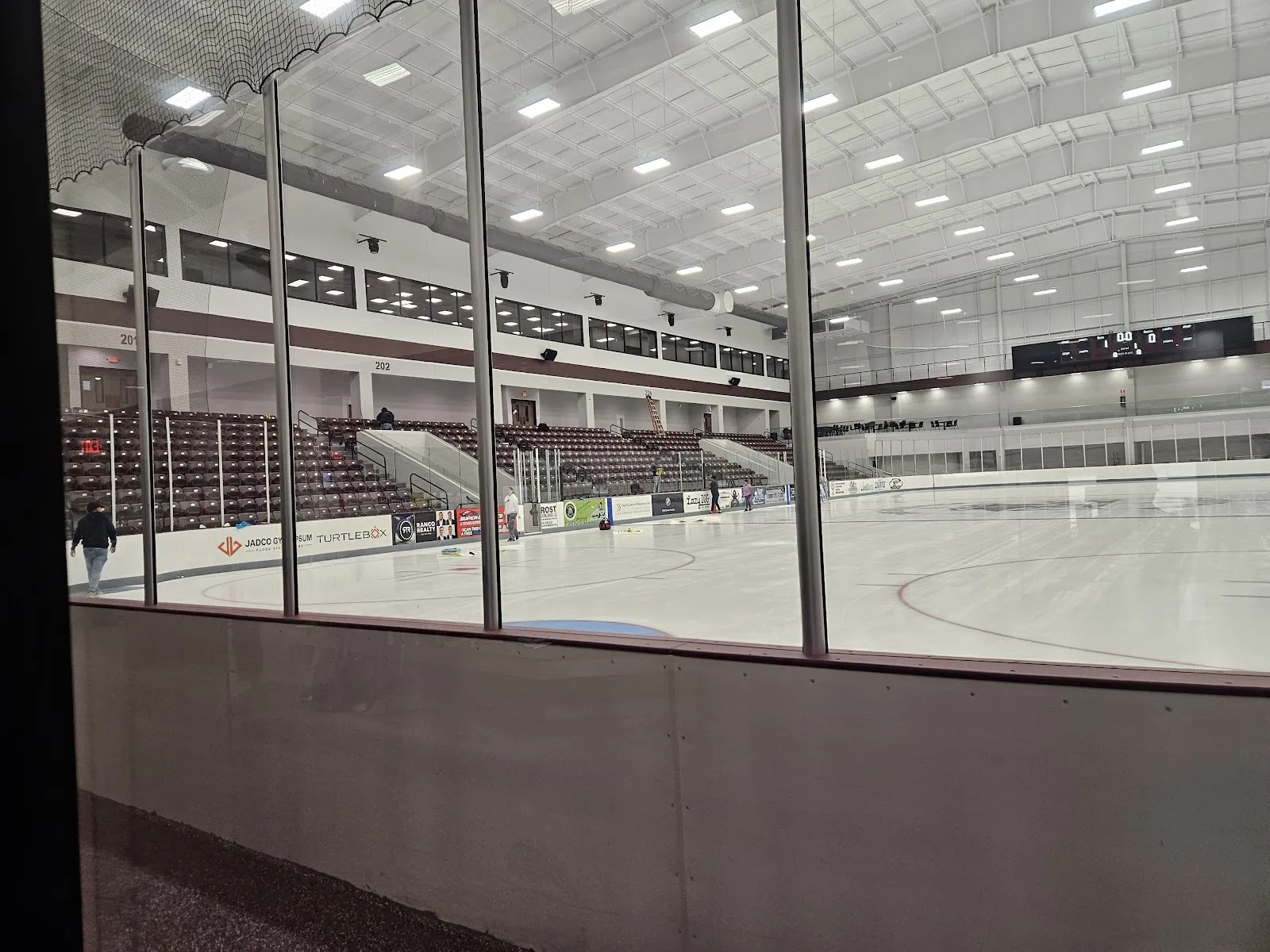 The empty sheet seen through dark glass from the concourse, white ice with a faint circle below tiered seating and a bright steel ceiling. — Deep South Ice and Sports Complex