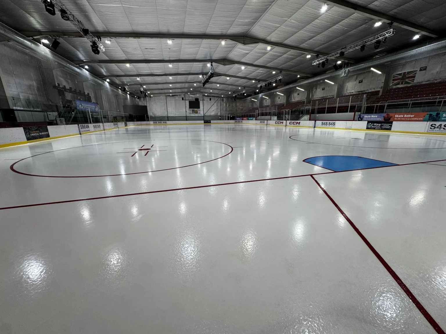 The empty Olympic-size ice from one corner, with the blue crease, red lines and the seating along the far side under the steel roof. — Deeside Ice Arena