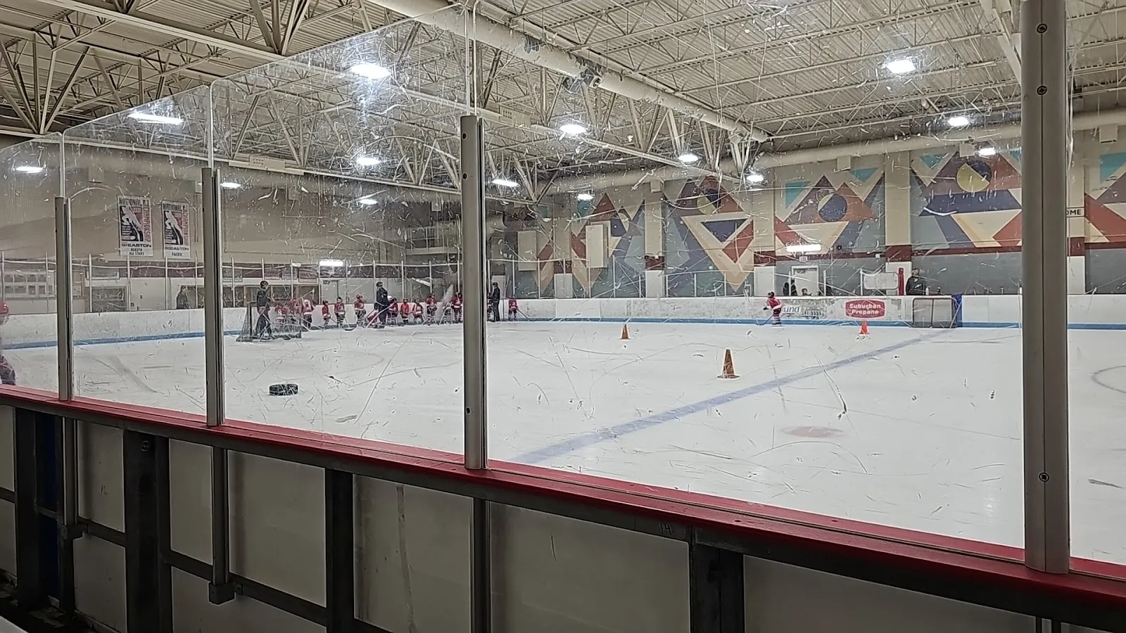The Dempsey Anderson ice seen past the boards, with a geometric mural along the far wall and a youth practice at the far end. — Dempsey Anderson Ice Arena