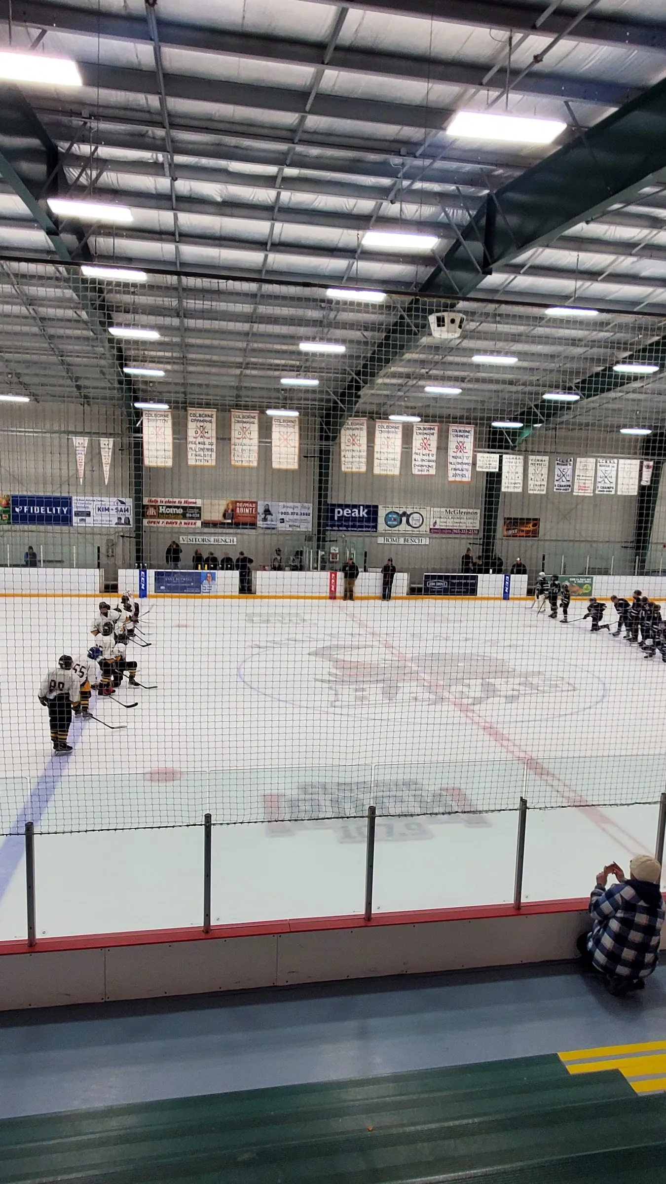 Una vista vertical de la pista con unos cuantos jugadores a lo lejos sobre el hielo, con una línea de vallas verdes y un techo de acero más allá. — Deseronto Community Centre