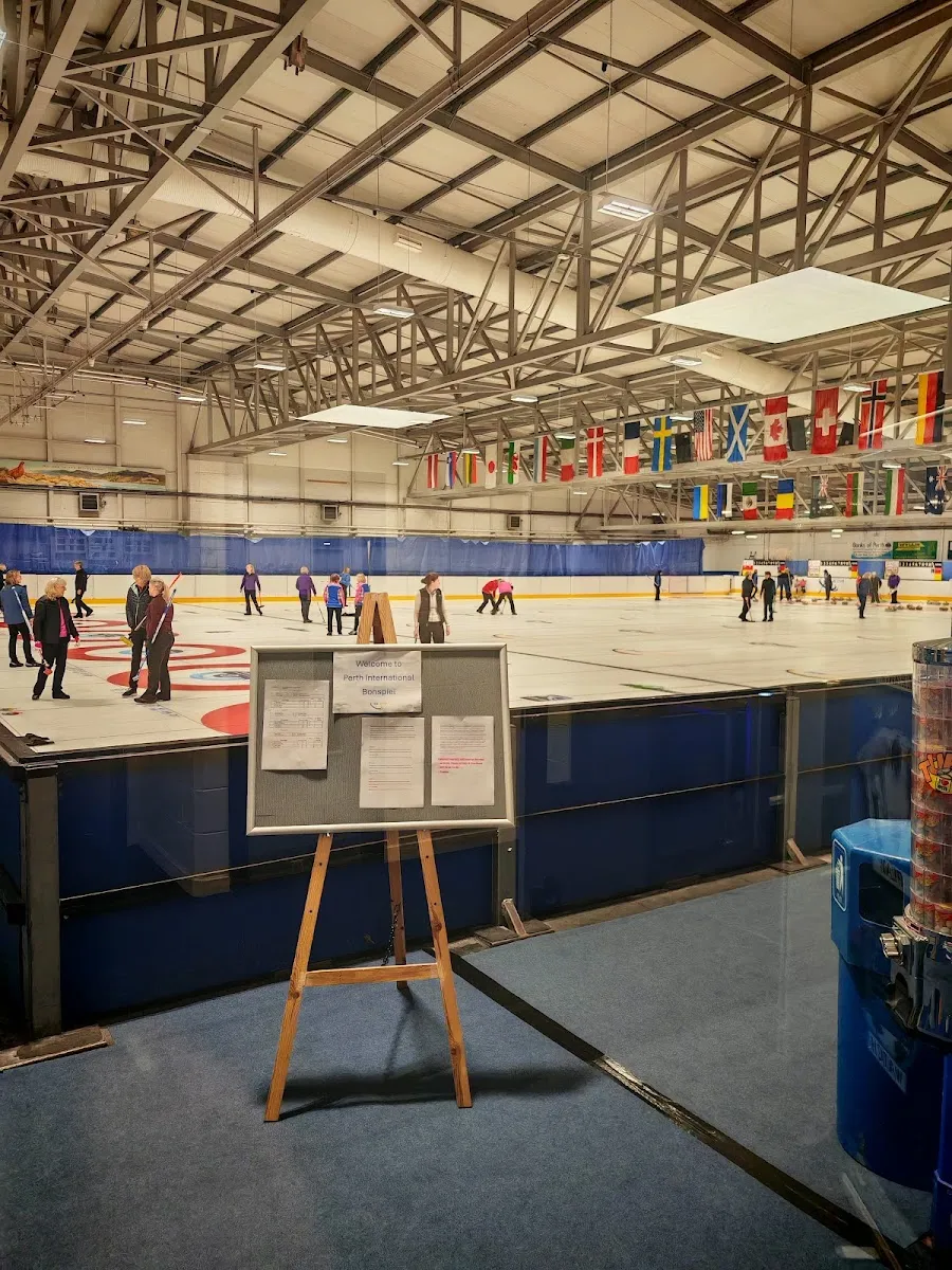 Curlers on the ice at Dewars Centre seen from the viewing rail, with a long row of national flags and the steel roof trusses above the far boards. — Dewars Centre