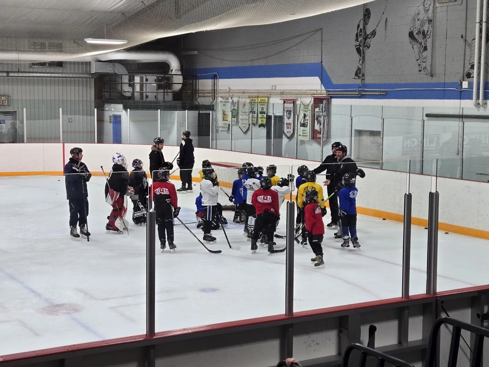 Jóvenes jugadores de hockey reunidos alrededor de sus entrenadores en el hielo durante una sesión en el Dickson Arena, con las vallas, el vidrio y los banderines de la pared detrás. — Dickson Arena
