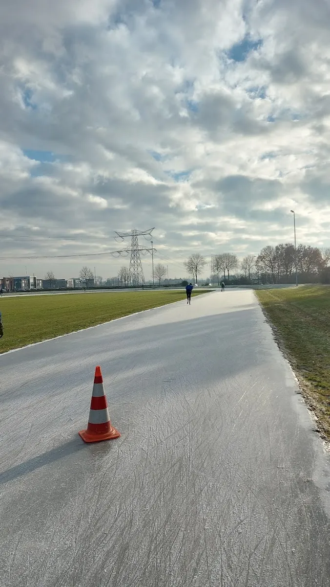 Skaters at a distance on the long natural-ice strip laid on the grass at the Dodewaardse IJsclub, with a traffic cone and pylons beyond – IJsbaan Dodewaard (Dodewaardse IJsclub)