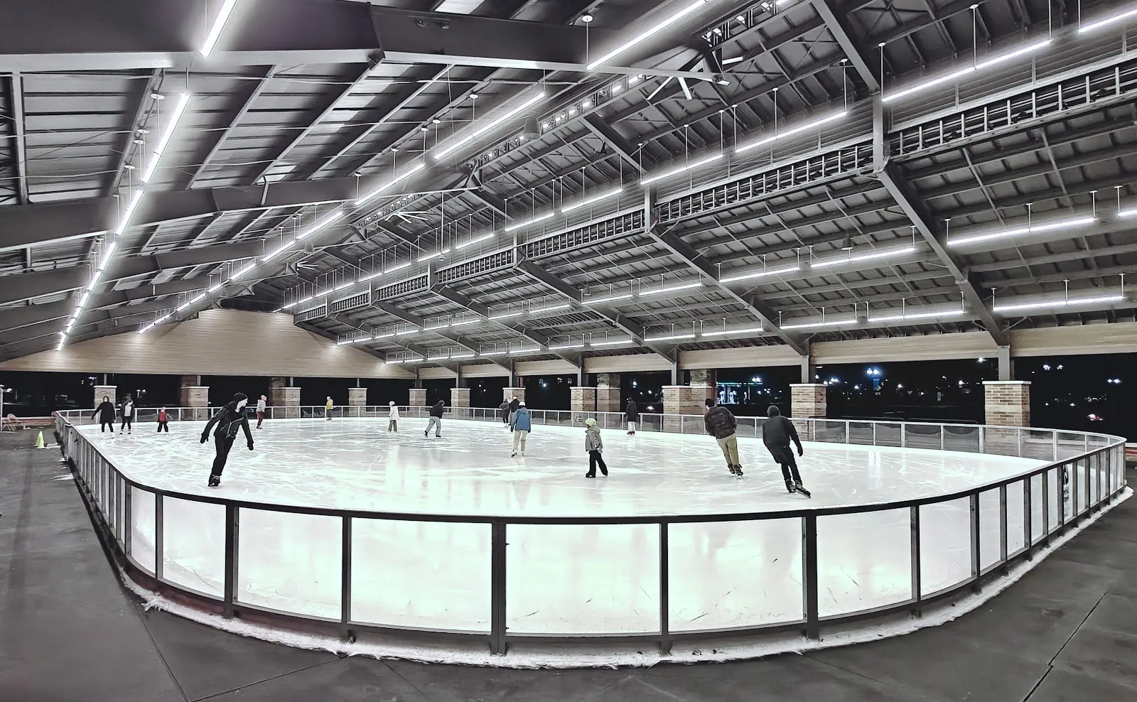 El pabellón techado y abierto sobre la pista refrigerada de noche, con las armaduras de acero del techo y la barandilla de vidrio. — Dodge Park Ice Rink