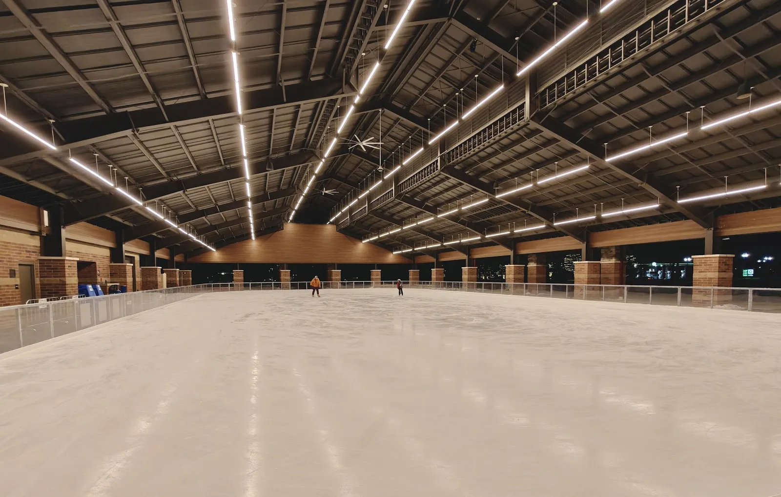 La pista vacía bajo el techo del pabellón, con pilares de ladrillo alrededor de la barandilla. — Dodge Park Ice Rink