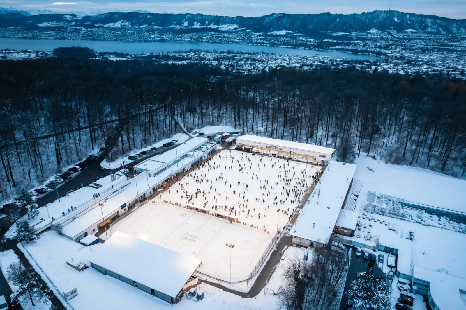 Aerial view of the Dolder Eisbahn on the Adlisberg in winter, the open ice surface between the grandstand building and the forest, with Lake Zurich beyond — Dolder Eisbahn
