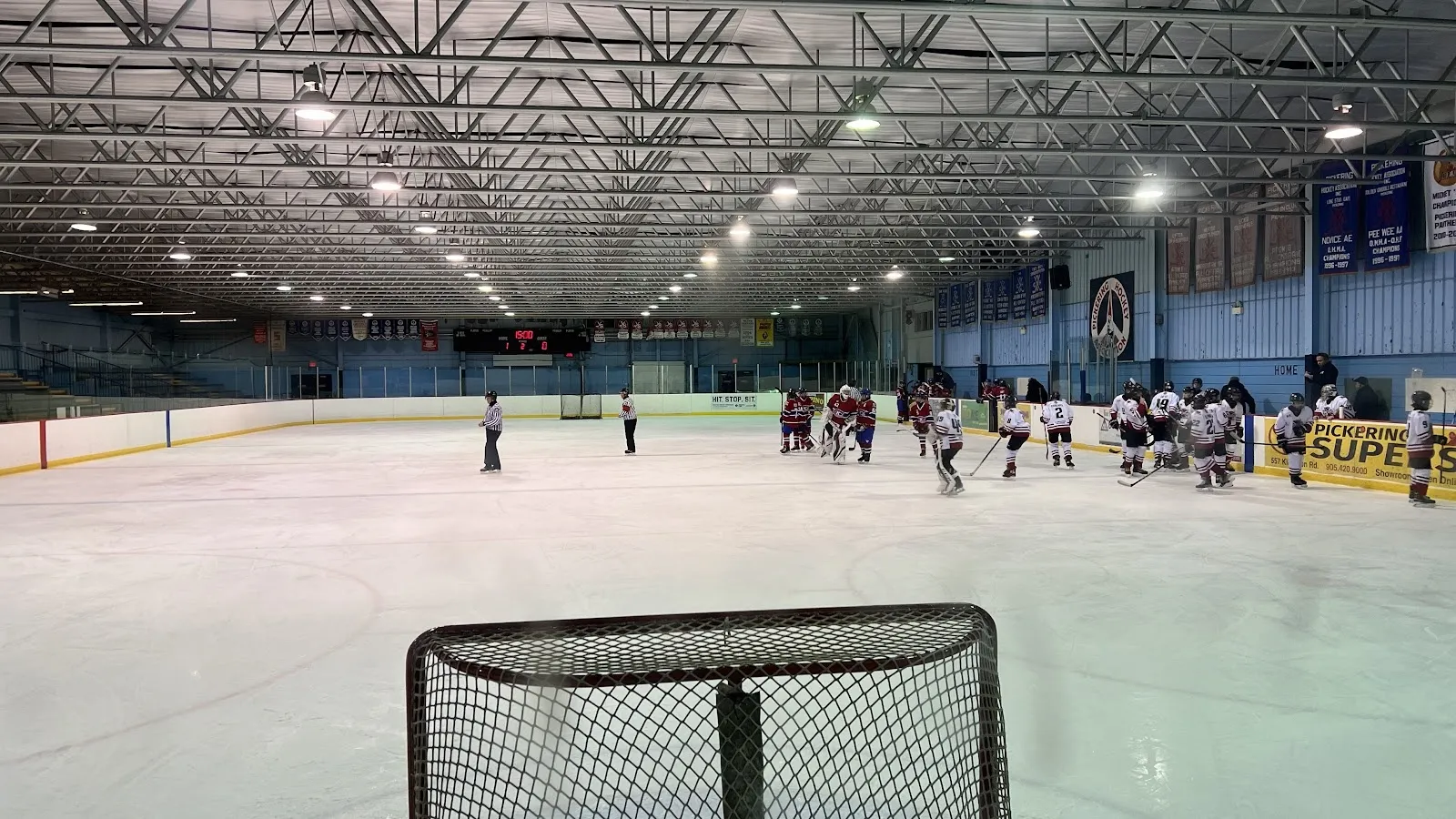 Una red en primer plano y un equipo practicando a lo ancho de una pista amplia bajo un oscuro techo de armaduras de acero, con banderines en la pared del fondo. — Don Beer Arena (Pickering)