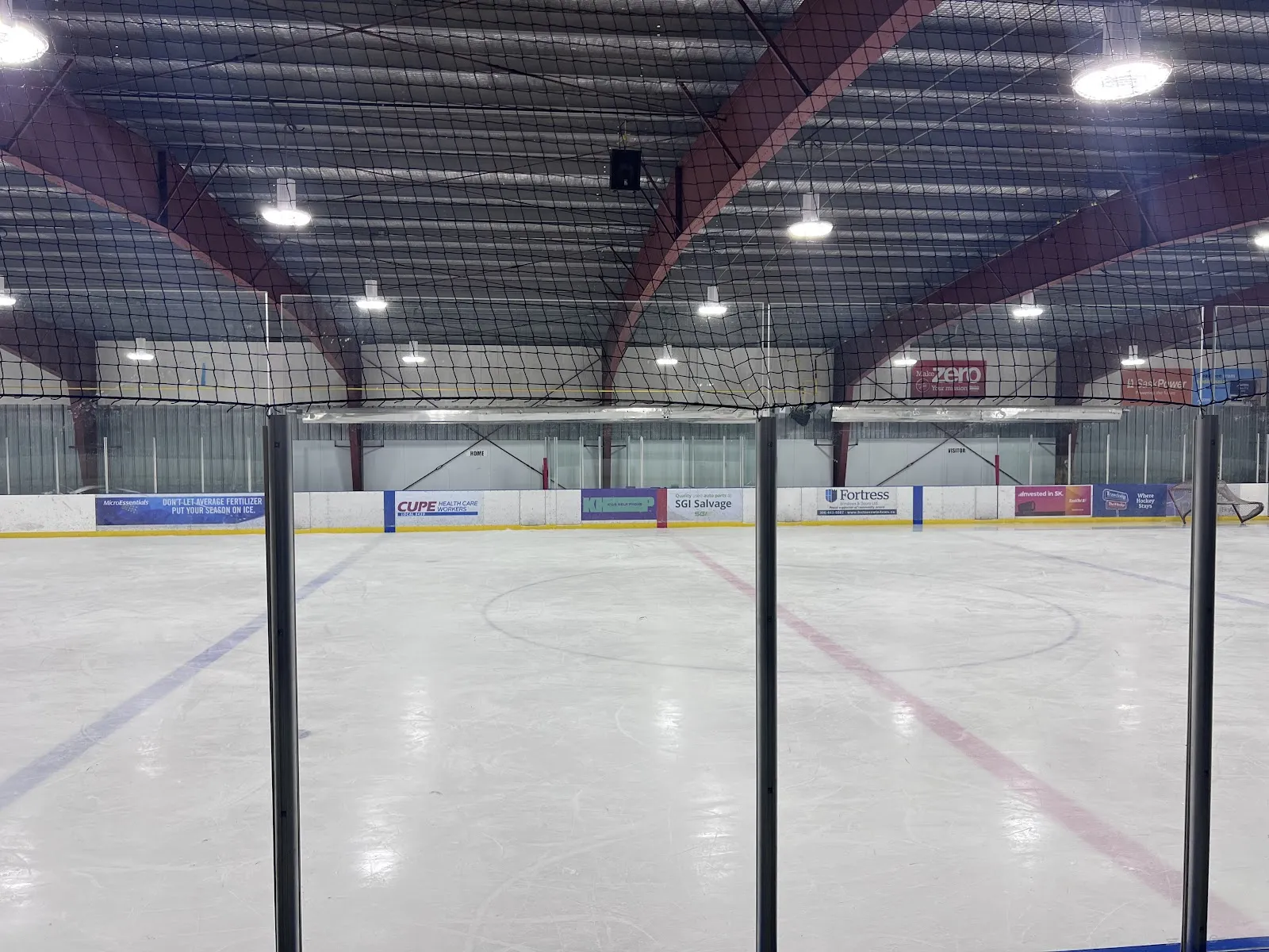 El hielo vacío de Don Ross Arena visto desde detrás de la red en un extremo, con la línea roja y anuncios de patrocinadores a lo largo del lado opuesto. — Don Ross Arena