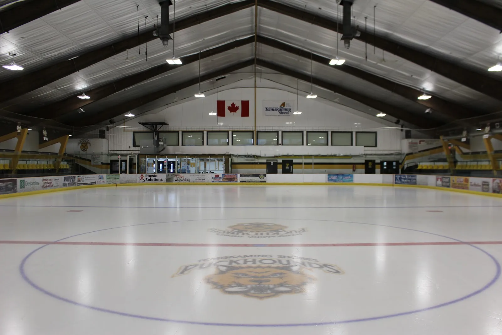 El hielo vacío del Don Shepherdson Memorial Arena con el logotipo de los Puckhounds al centro, bajo un techo de madera a dos aguas. — Don Shepherdson Memorial Arena (New Liskeard)