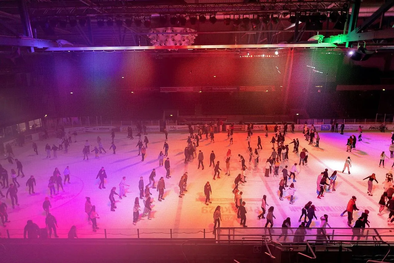Public skating night in the Donau-Arena: skaters spread across the whole ice sheet under the steel roof, lit in red and orange, with the video cube above centre ice. — das Stadtwerk.Donau-Arena