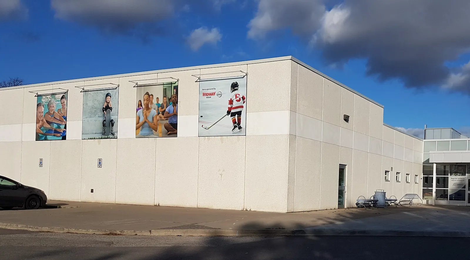 A side wall of the complex in white block with four large banner photographs of swimmers and a young hockey player, and the glazed entrance at right. — Donevan Recreation Complex