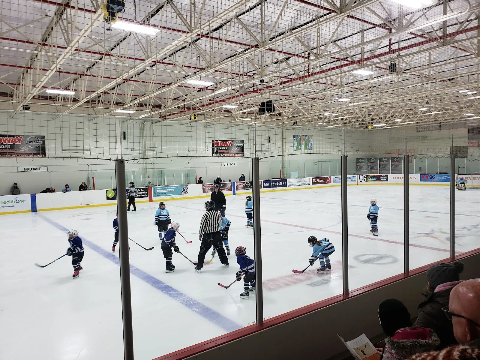 Young players skating a hockey practice on the ice at Donevan Recreation Complex, seen through the glass from the stands, with the boards and steel roof trusses above. — Donevan Recreation Complex