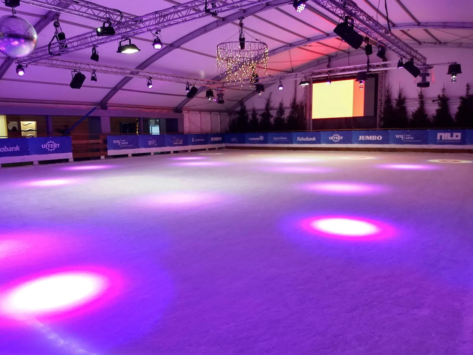 The empty tented ice rink of DongenIce lit in purple, seen from the boards with the sponsor banners behind – DongenIce (Dongen)