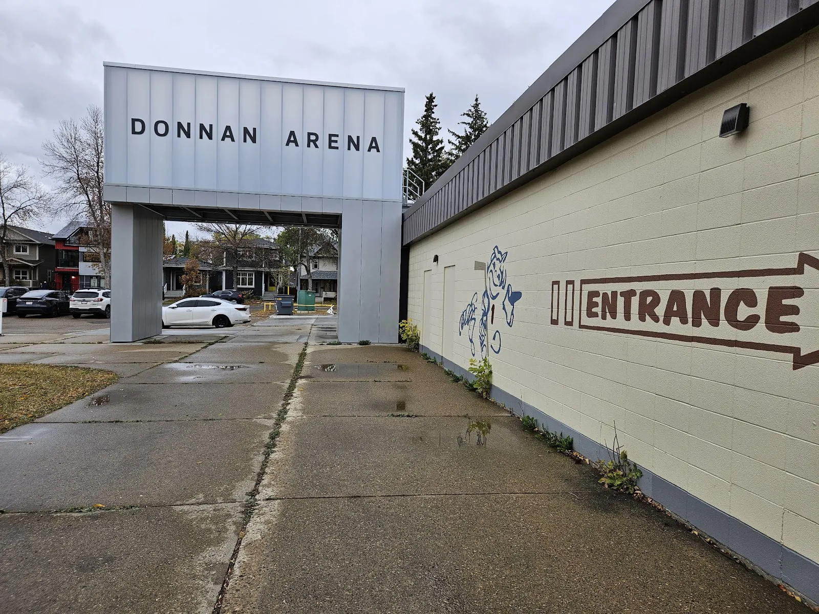Grey metal building entrance canopy reading "DONNAN ARENA" over a walkway, with a side entrance marked "ENTRANCE" on the beige cinderblock wall of the rink building, parked cars and houses visible beyond. — Donnan Arena