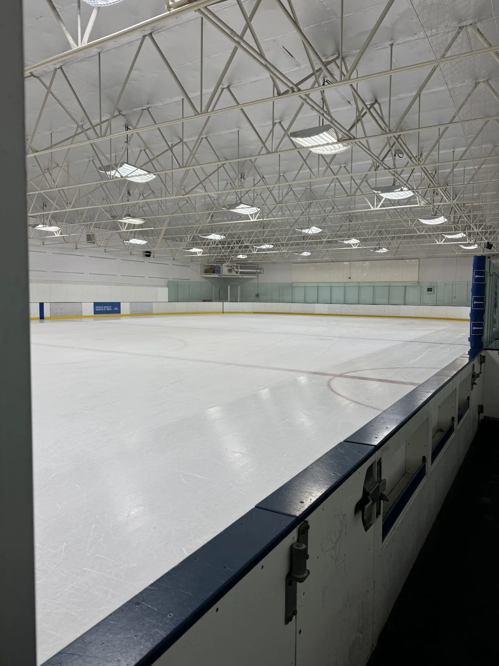 Empty single-sheet ice rink seen from the boards, showing the full ice surface, glass and board along the far side, exposed white steel roof trusses overhead, and a blue "Edmonton" board ad in the corner. — Donnan Arena