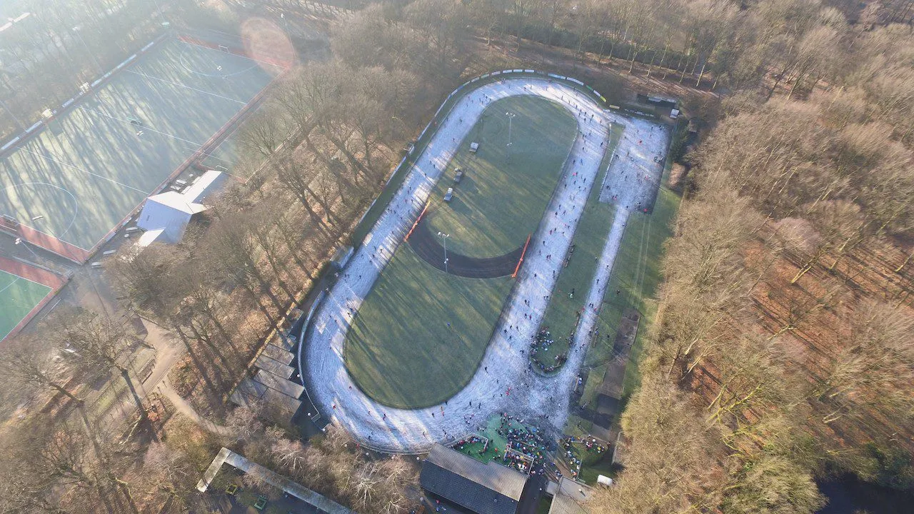 Luchtfoto van de ovale baan met ijs, omgeven door bos, met de daken van het clubhuis onderaan. – Doornsche IJsclub (Doorn)