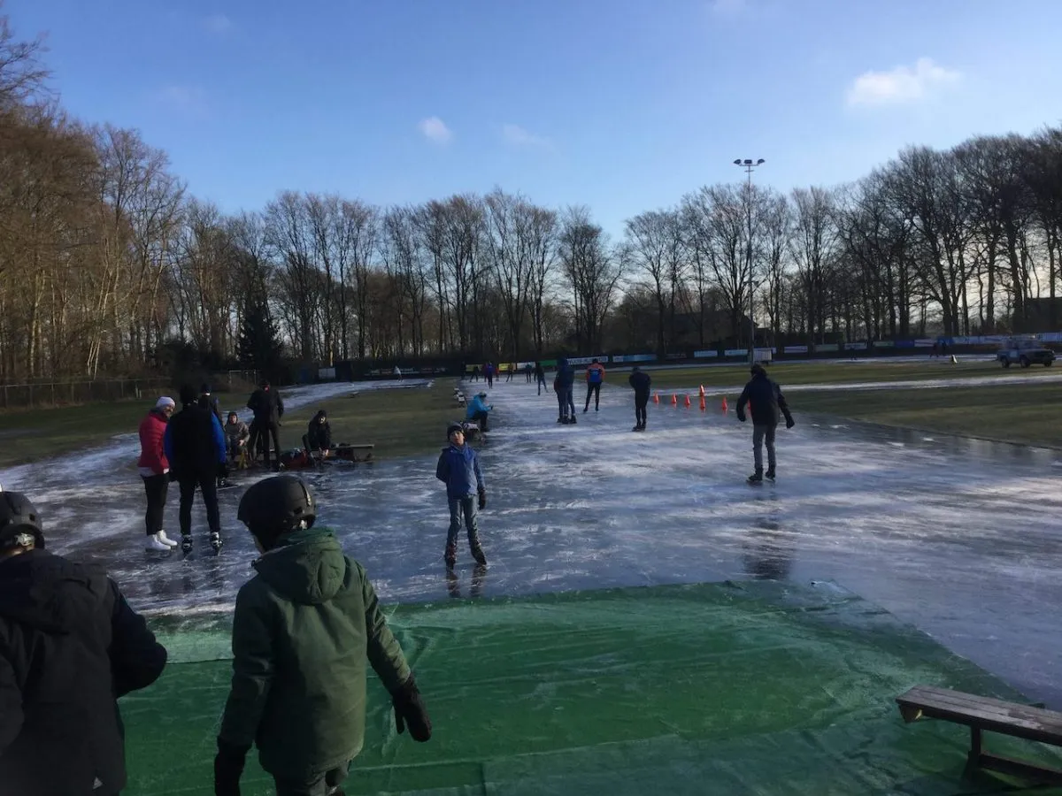 Schaatsers verspreid over het vers gemaakte ijs van de Doornse baan, met pylonen, een bank en hoge kale bomen erachter onder een blauwe lucht. – Doornsche IJsclub (Doorn)