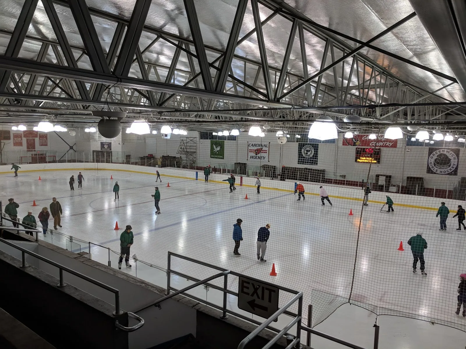 Una sesión pública en una pista amplia, con patinadores repartidos entre conos naranjas bajo un oscuro techo con refuerzos de acero y banderines en la pared del fondo. — Dorothy Hamill Ice Rink