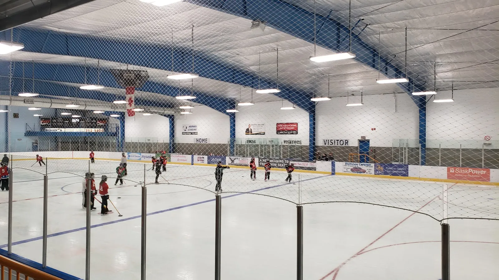 Interior view of the full ice sheet from the stands during a minor-hockey practice, showing the boards, netting, a Canadian flag, and HOME/VISITOR bench signage. — Doug Wickenheiser Arena