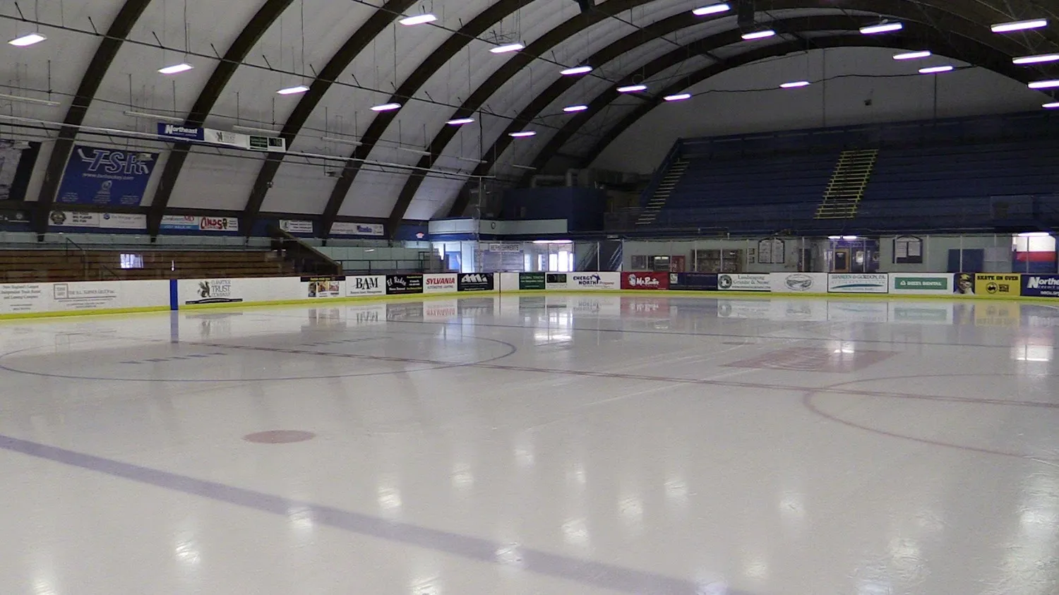 Toda la pista vacía de Douglas N. Everett Arena bajo un techo en arco de costillas de madera, con gradas azules escalonadas y una fila de anuncios de patrocinadores locales a lo largo de las vallas. — Douglas N. Everett Arena