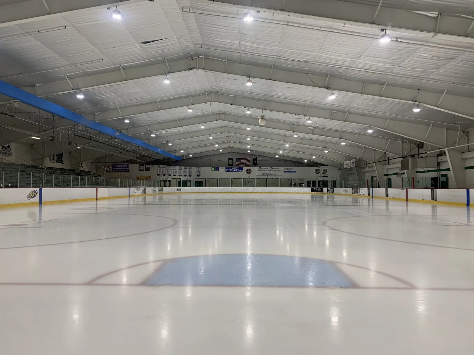 Wide view of the empty ice sheet from ice level, looking down the rink toward the far boards with championship banners and an American flag hanging above. — Dover Ice Arena