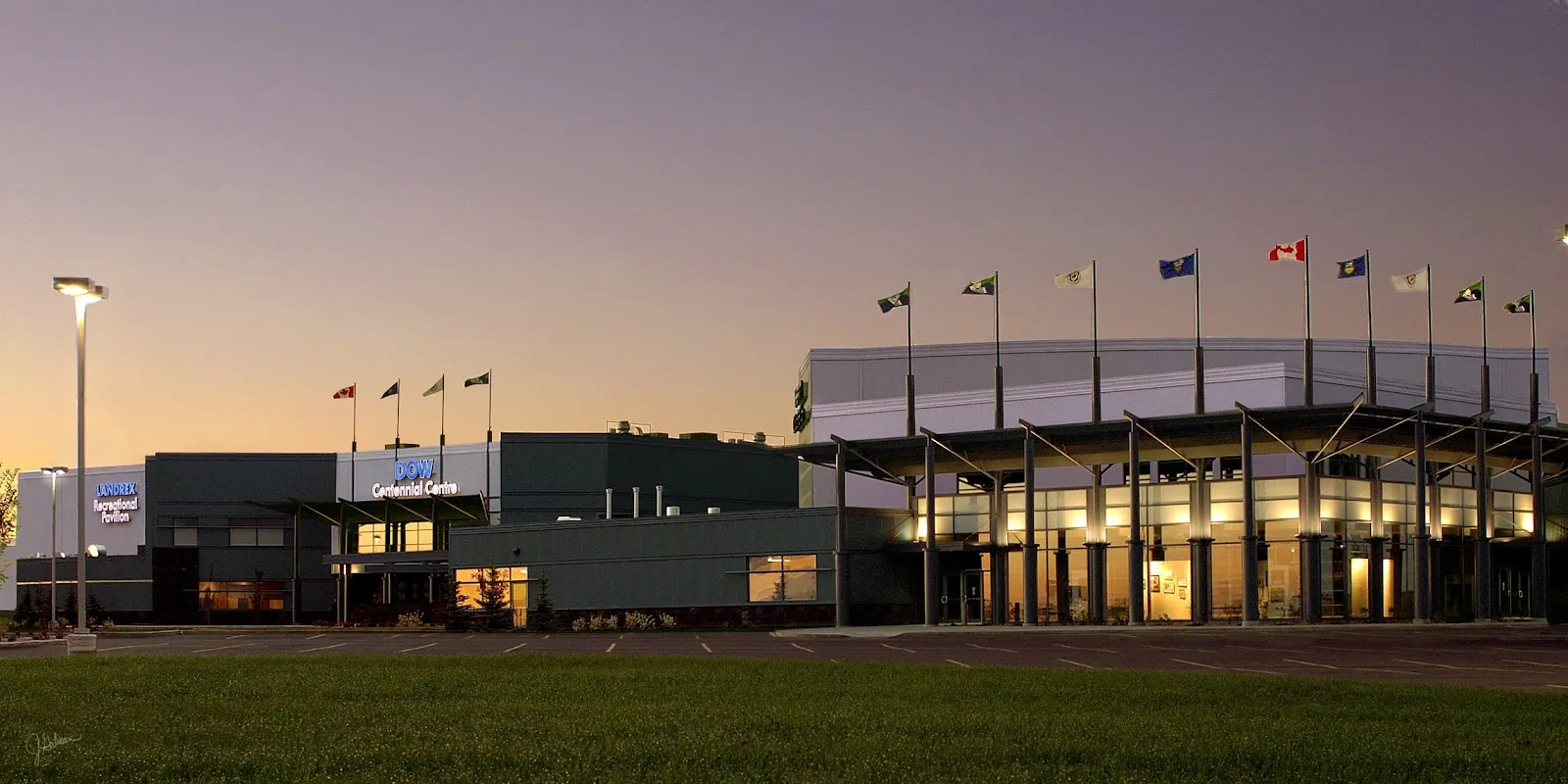 The Dow Centennial Centre at dusk: lit glass and a row of flags along the roofline above the main entrance. — Dow Centennial Centre