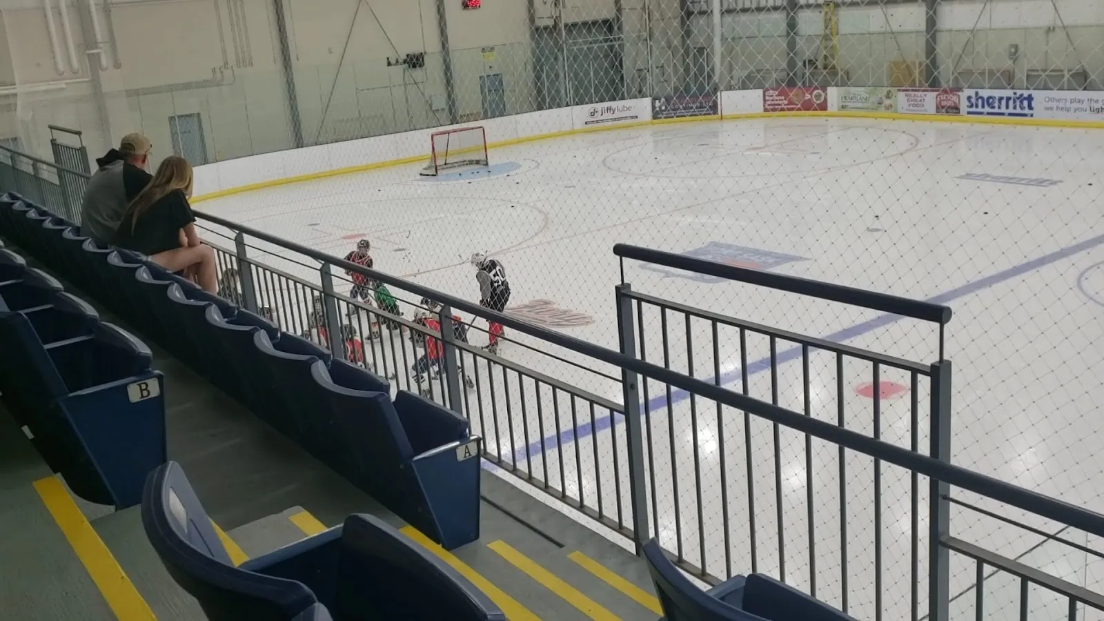 A wide view of the sheet from behind the glass, the ice empty with a few people at the boards and a banner of yellow and white seats along the near side. — Dow Centennial Centre