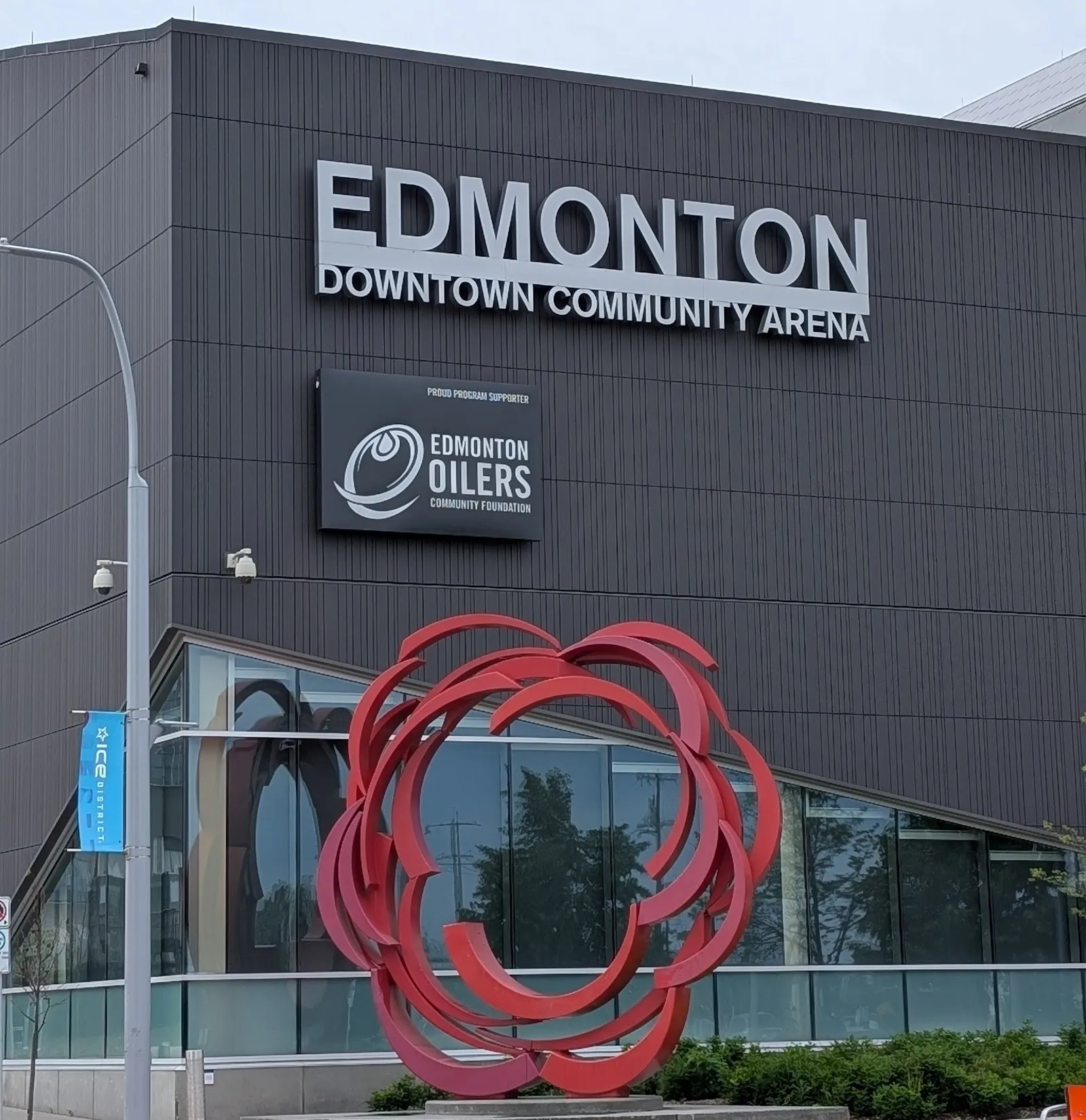 La Downtown Community Arena de Edmonton, con revestimiento oscuro y su nombre en letras en lo alto del muro, sobre una escultura de listón rojo — Downtown Community Arena