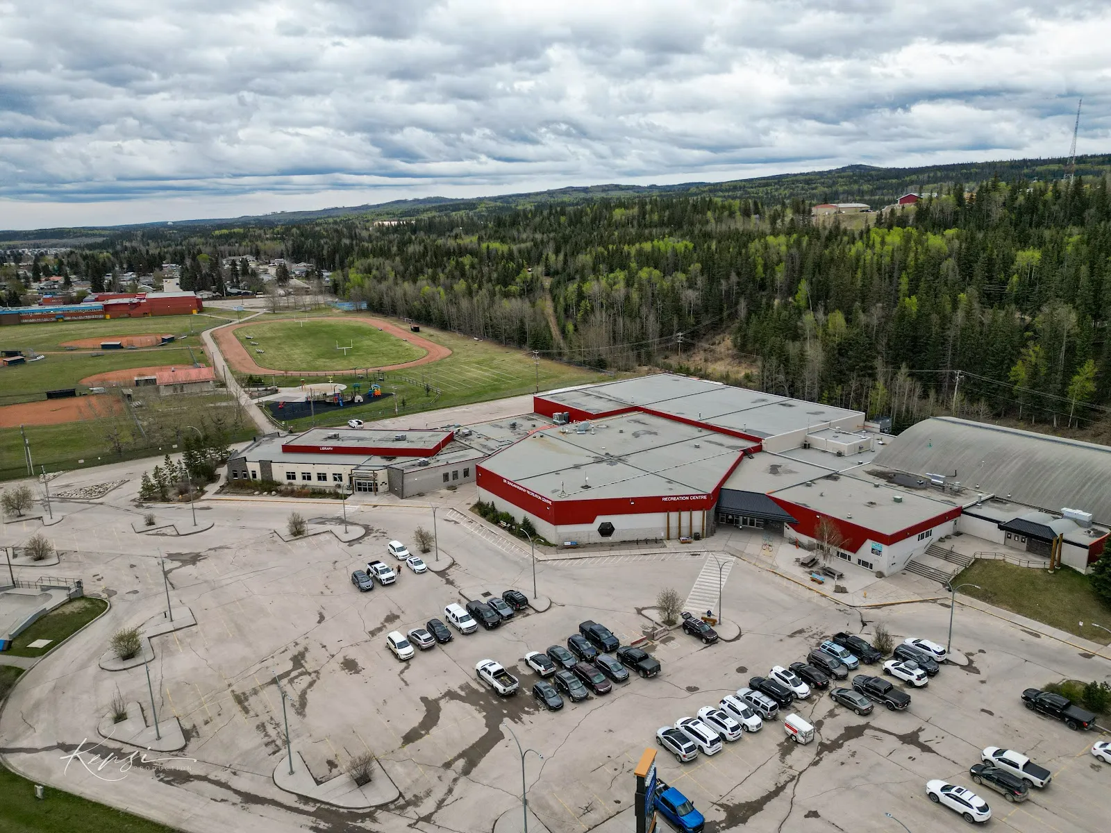 An aerial view of the Dr. Duncan Murray Recreation Centre in Hinton, with its red-and-grey roofs, the parking lot in front and ball diamonds and forested hills behind. — Dr. Duncan Murray Recreation Centre