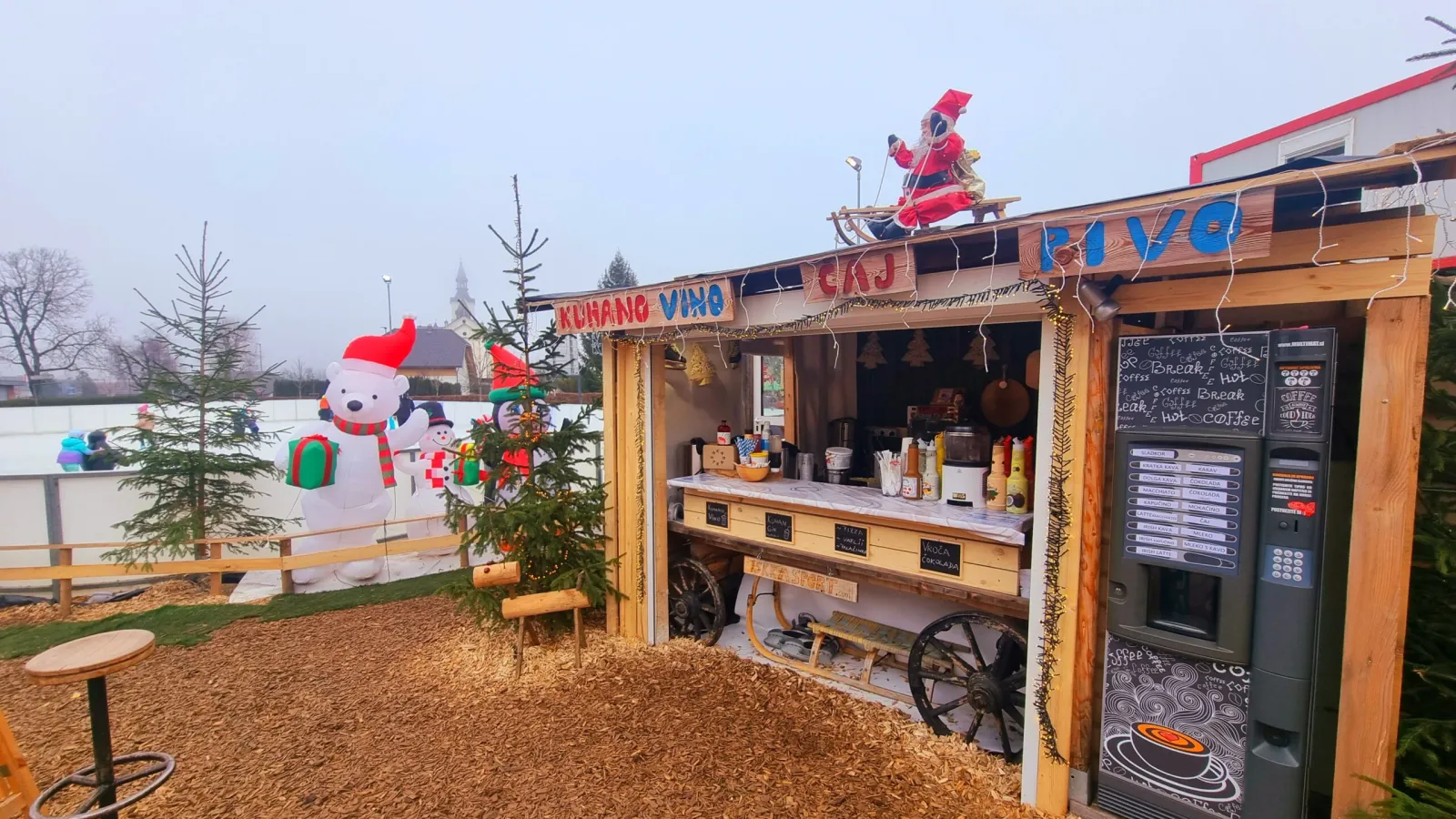 The wooden bar hut at the Bohinjska Bistrica rink at dusk, with mulled wine, tea and beer signs, a coffee machine and festive inflatables, and the ice behind it. — Drsališče Bohinjska Bistrica