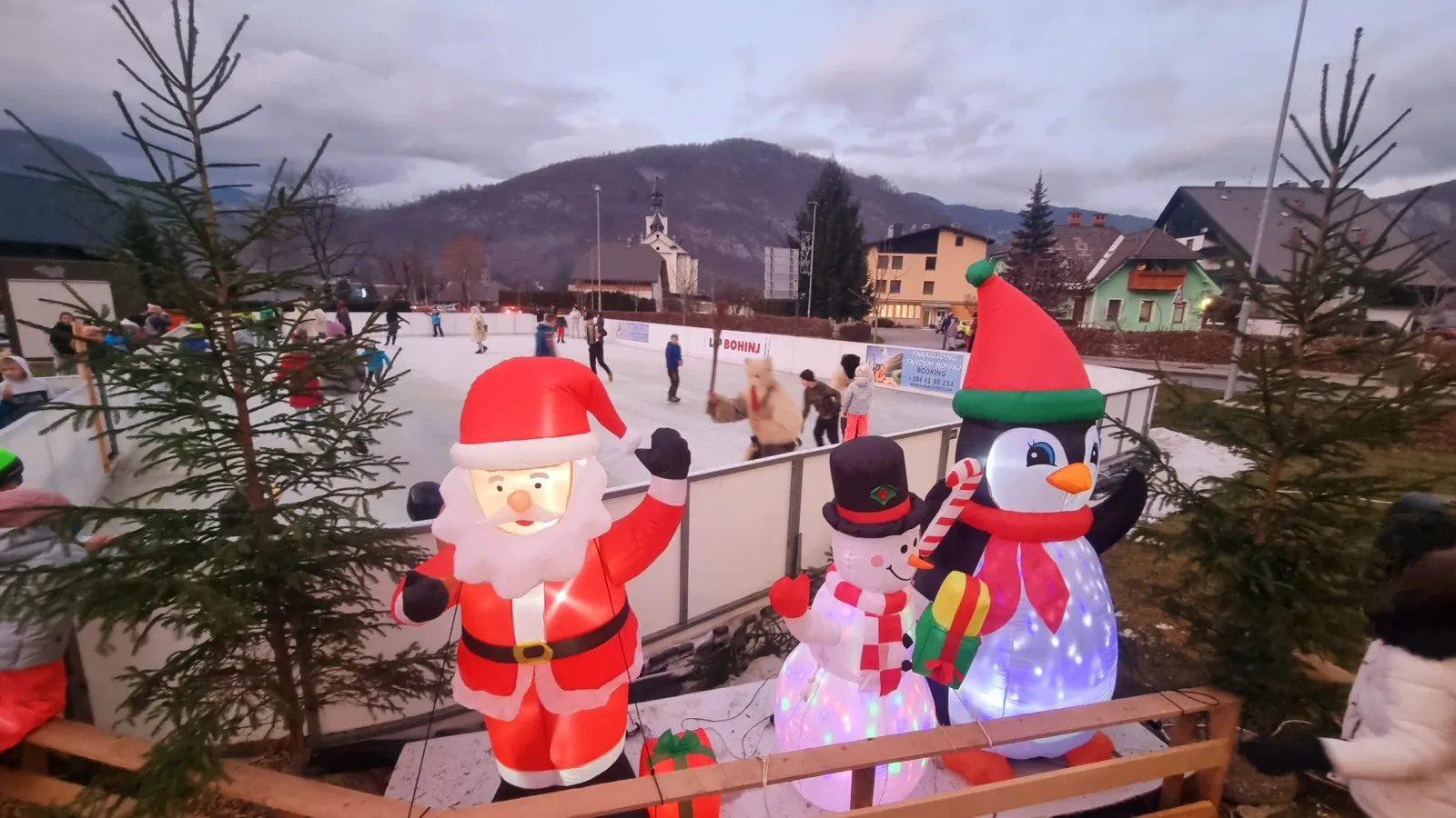 Skaters on the white ice of the Bohinjska Bistrica rink, with festive inflatable figures at the boards and the mountains and village rooftops behind. — Drsališče Bohinjska Bistrica