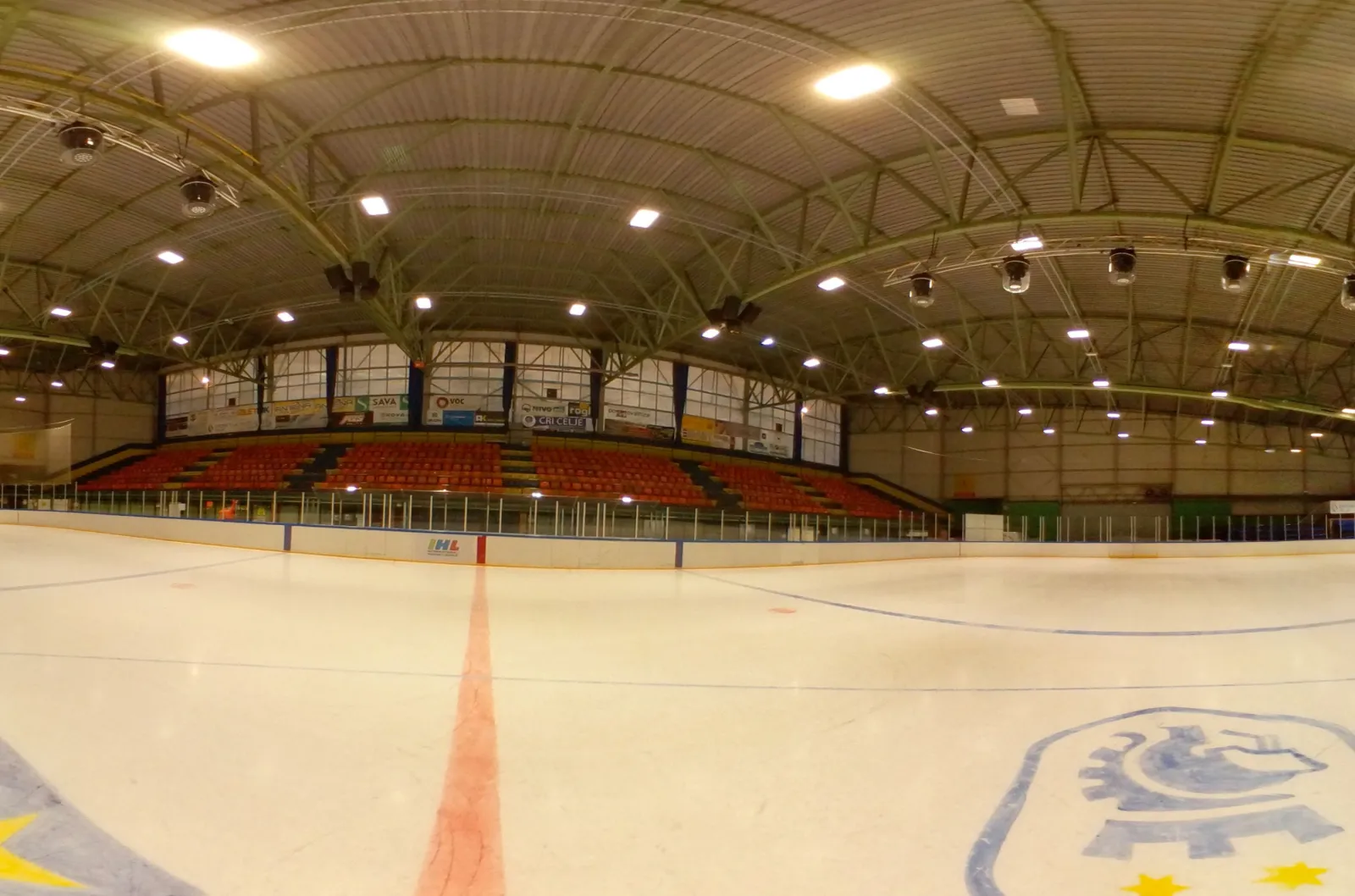 The empty ice sheet of the Drsališče v Mestnem parku in Celje seen from the boards, with the red-seated stand, the steel-trussed roof and the centre line, cropped from the operator's 360-degree photo — Drsališče v Mestnem parku Celje