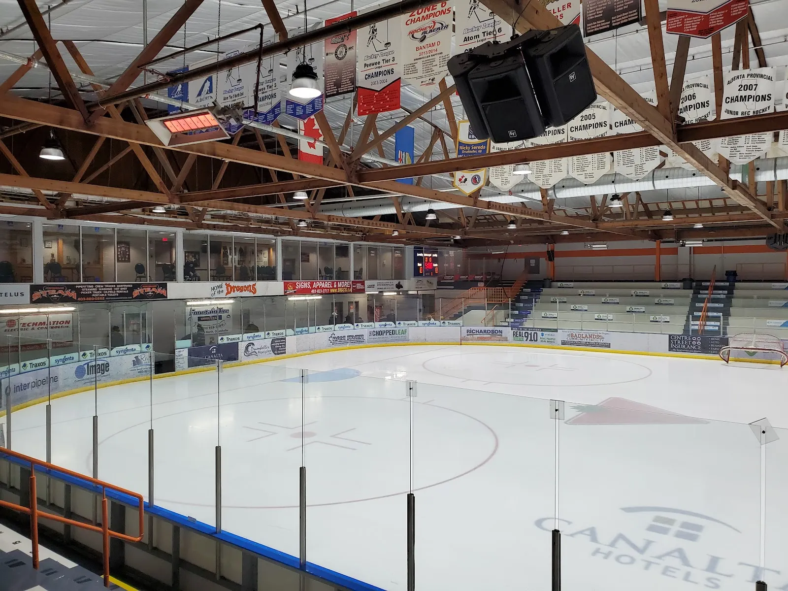 The empty ice at the Drumheller Memorial Arena under timber trusses hung with championship banners, with the Dragons' boards and stands beyond. — Drumheller Memorial Arena