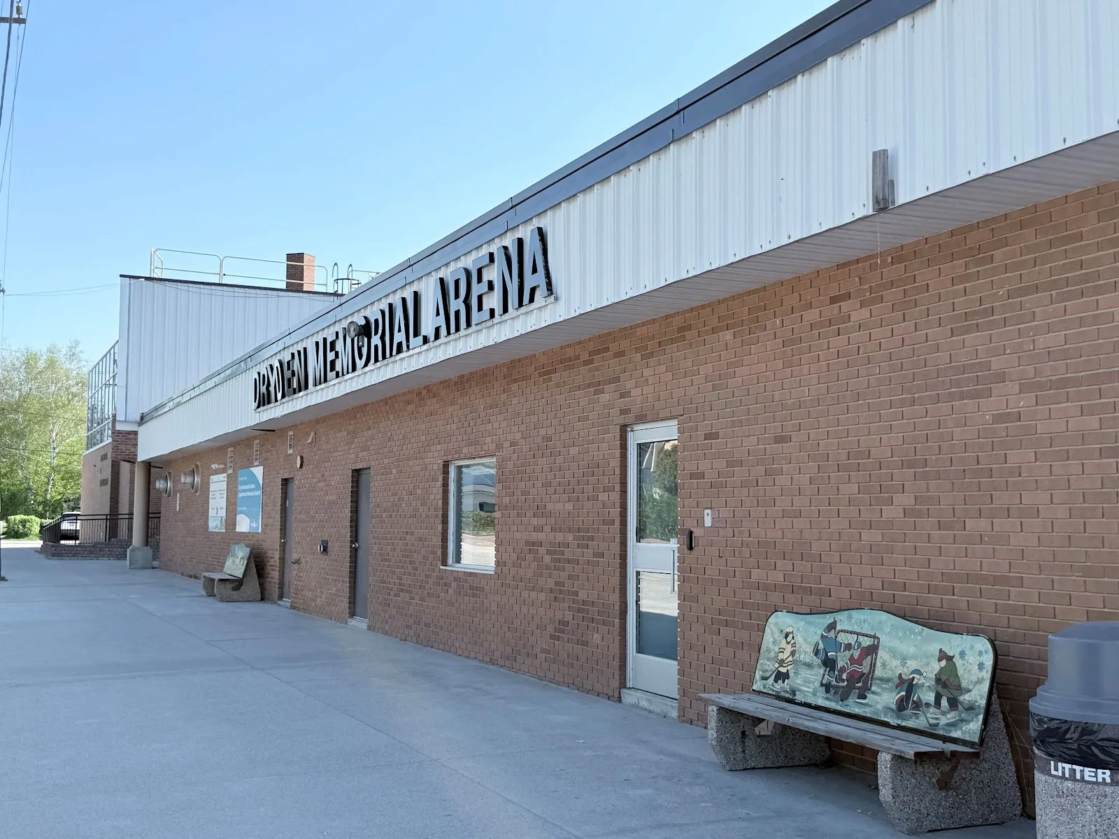 The brick Dryden Memorial Arena side wall with its name above the door and a bench in front. — Dryden Memorial Arena