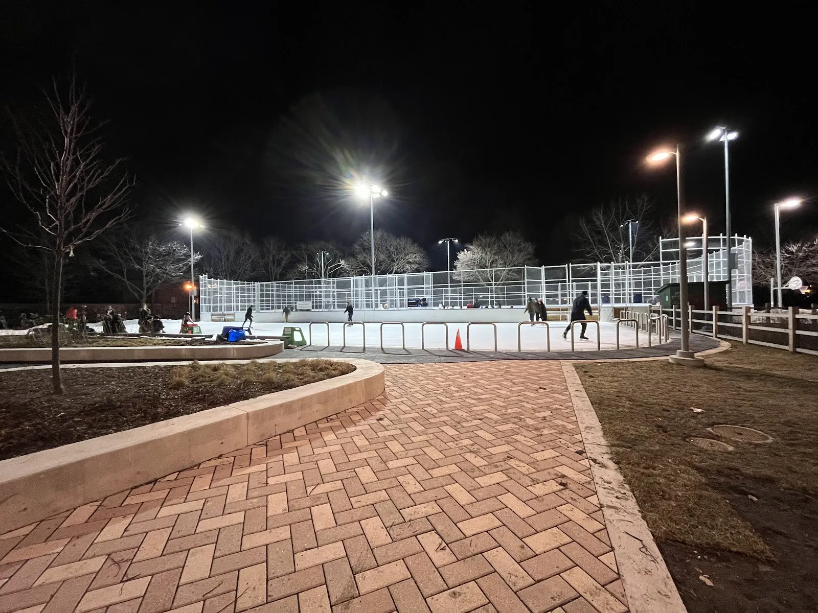 The lit, fenced outdoor rink at Dufferin Grove Park at night in winter seen from the brick path, with a few skaters far off on the ice — Dufferin Grove Park Outdoor Ice Rinks