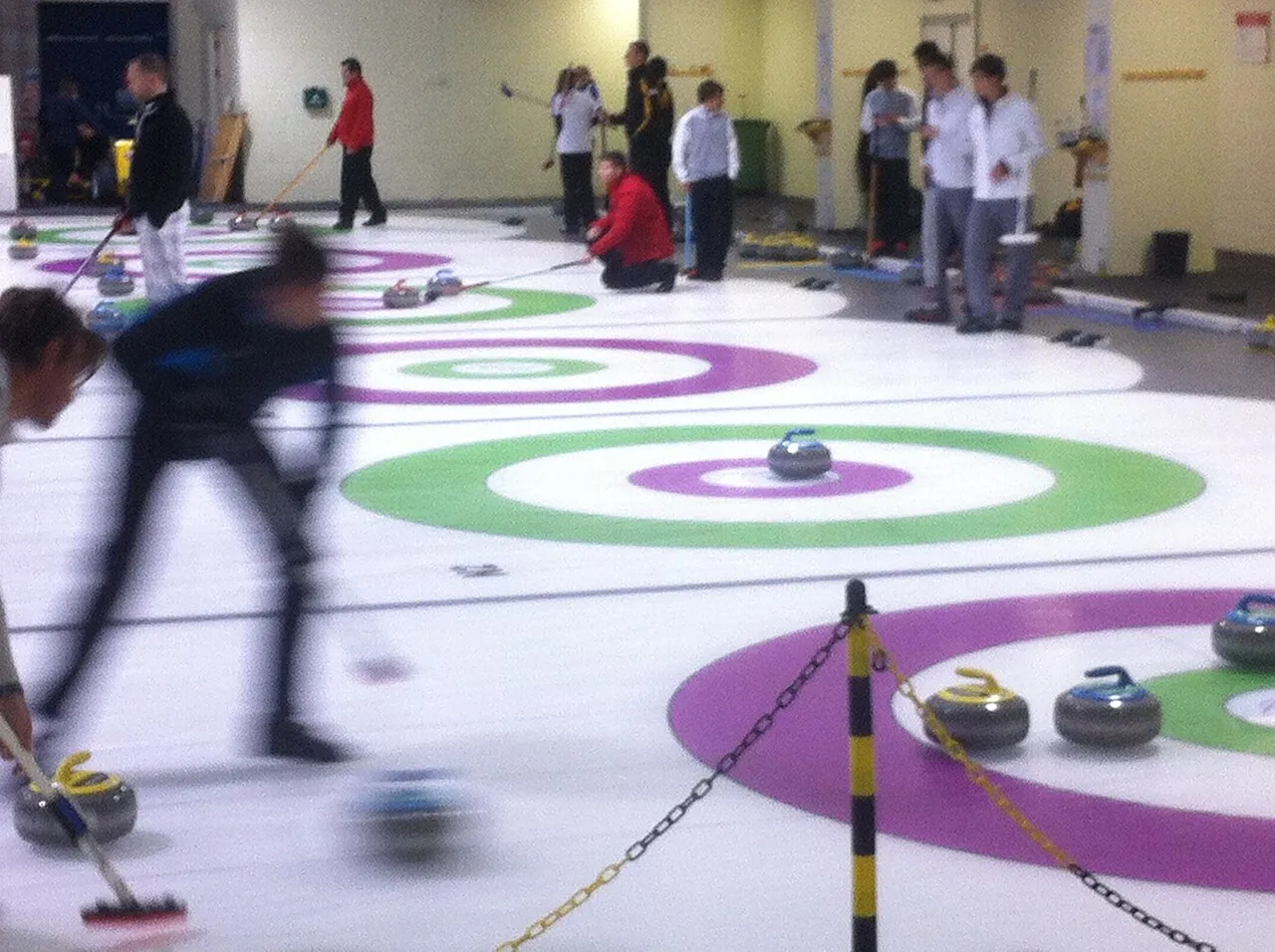 Curlers sweeping on the ice pad at the Dumfries Ice Bowl, with stones and the painted houses in the foreground — Dumfries Ice Bowl