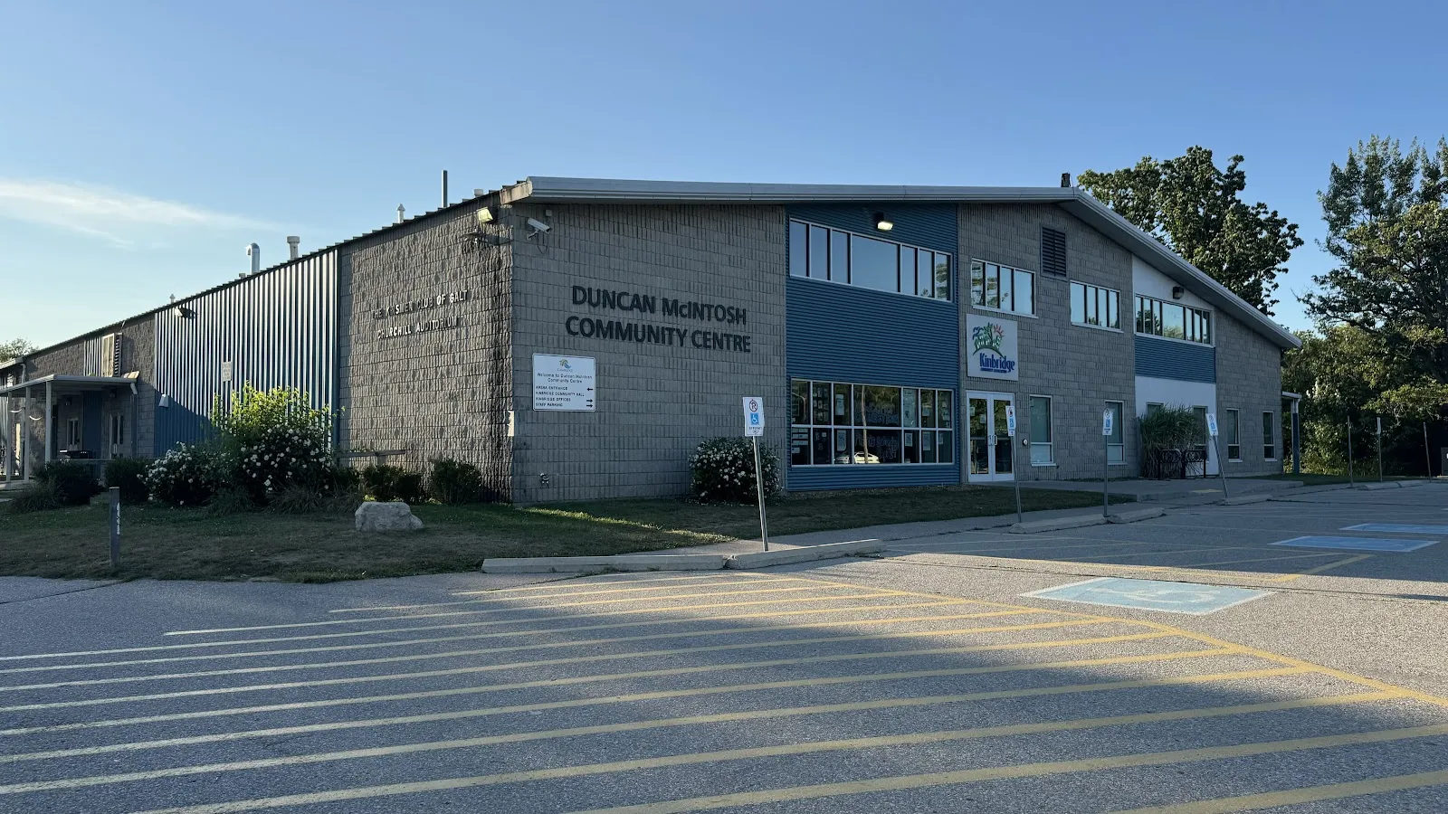 El Duncan McIntosh Community Centre, un edificio de dos plantas de ladrillo y paneles azules con su nombre en la fachada, visto al otro lado de un aparcamiento con rayas bajo un cielo azul despejado. — Duncan McIntosh Arena