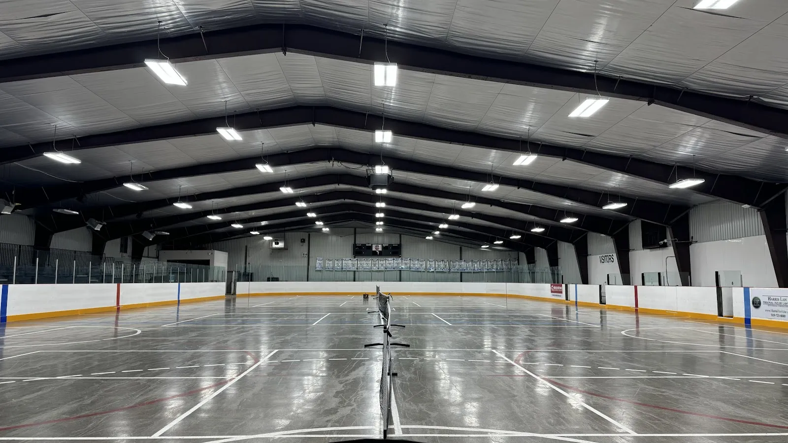 The empty rink from centre ice, with a net at centre, white boards and a dark arched roof with rows of lights. — Duncan McIntosh Arena