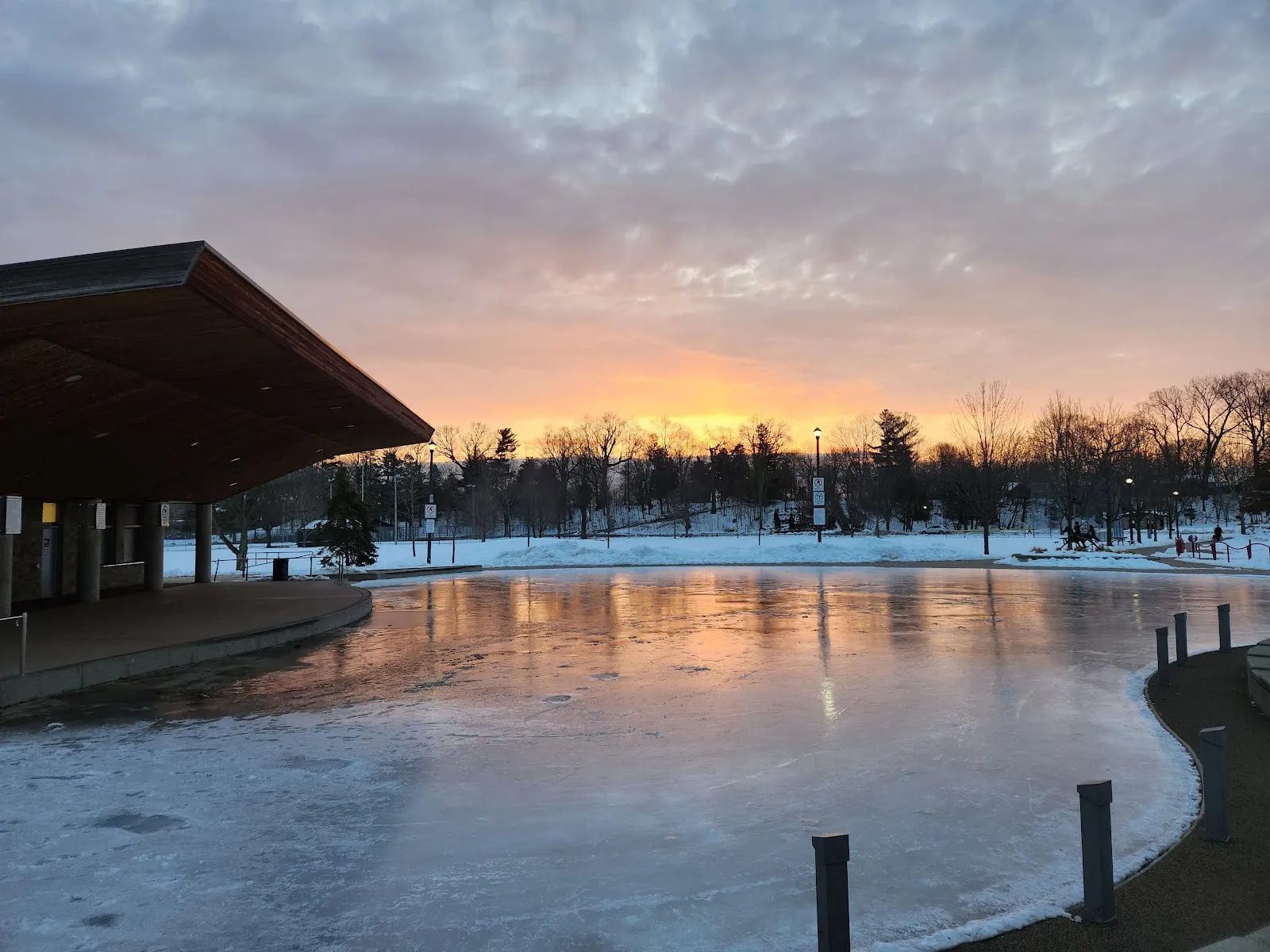 La pista estilo estanque del Dundas Driving Park al atardecer, con la superficie de hielo junto al pabellón del parque, nieve y árboles al fondo y sin personas a la vista — Dundas Driving Park City Outdoor Rink