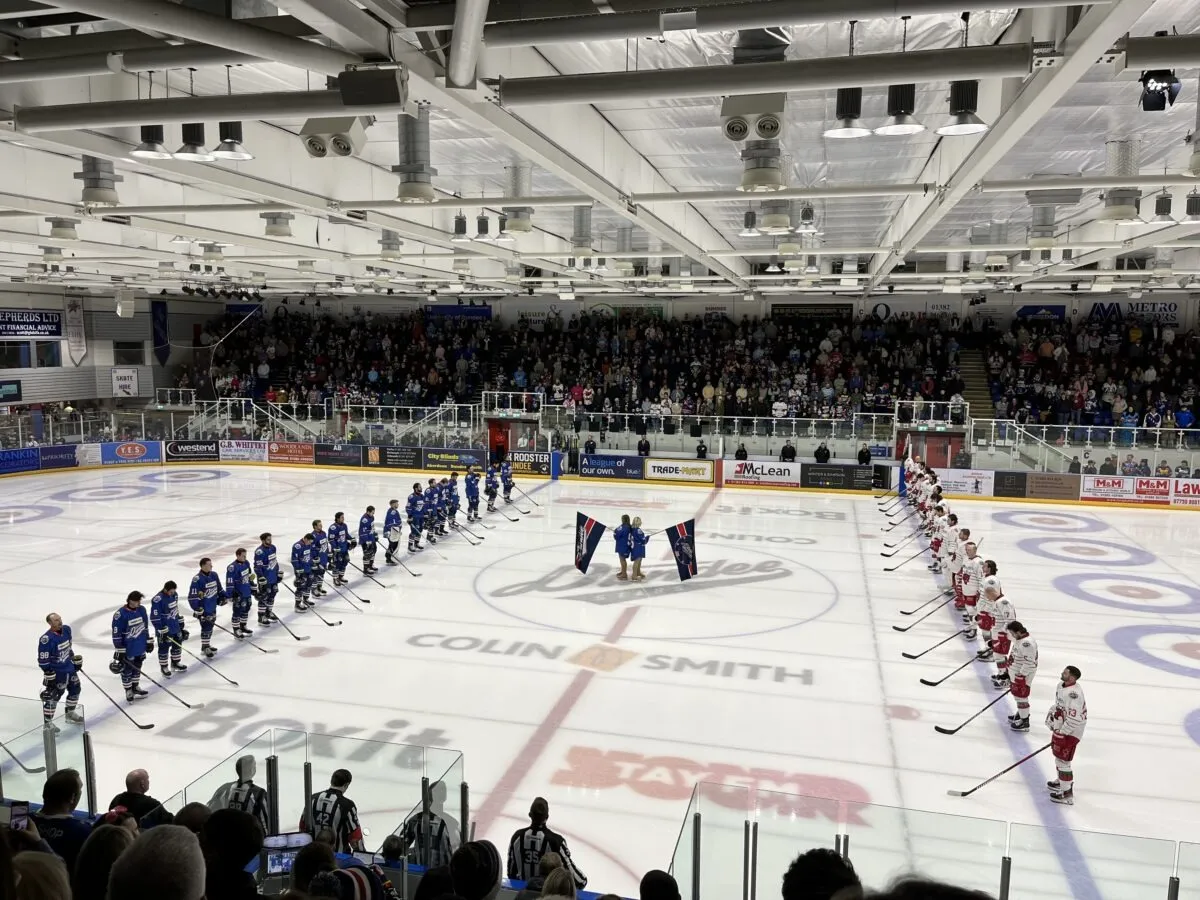 The arena before a Dundee Stars home game, both teams lined up on the blue lines on the ice with the stands full behind them and the centre-ice logo in view. — Dundee Ice Arena