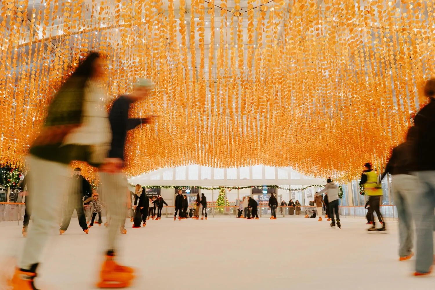 Skaters gliding on the ice beneath a canopy of hanging amber lights at Dundrum On Ice, other skaters in the tented rink beyond. — Dundrum On Ice