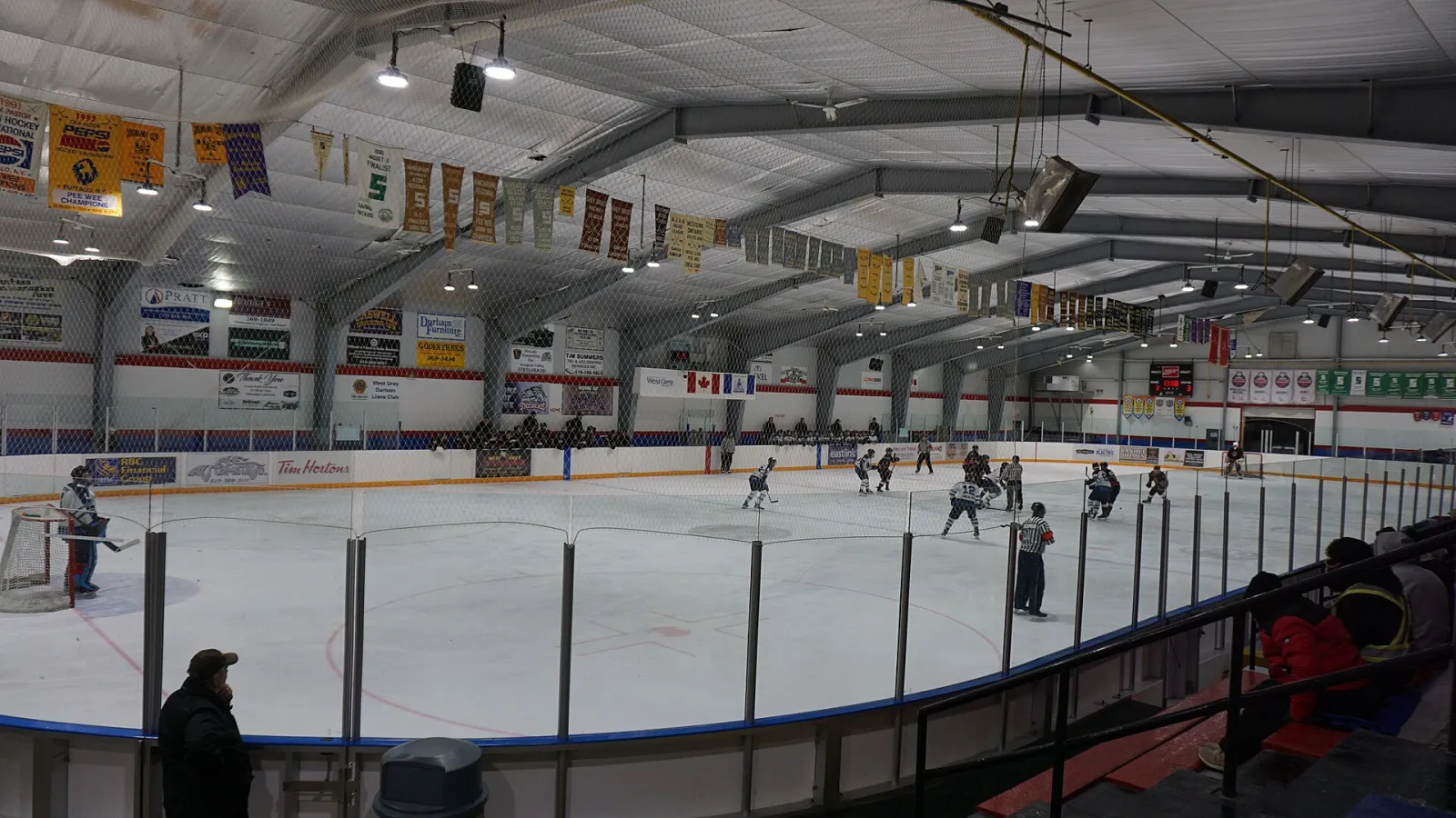 El hielo del Durham Community Centre durante un partido de hockey en febrero de 2026, visto desde detrás del vidrio bajo el techo arqueado de acero con estandartes de campeonatos. — Durham Community Centre and Arena
