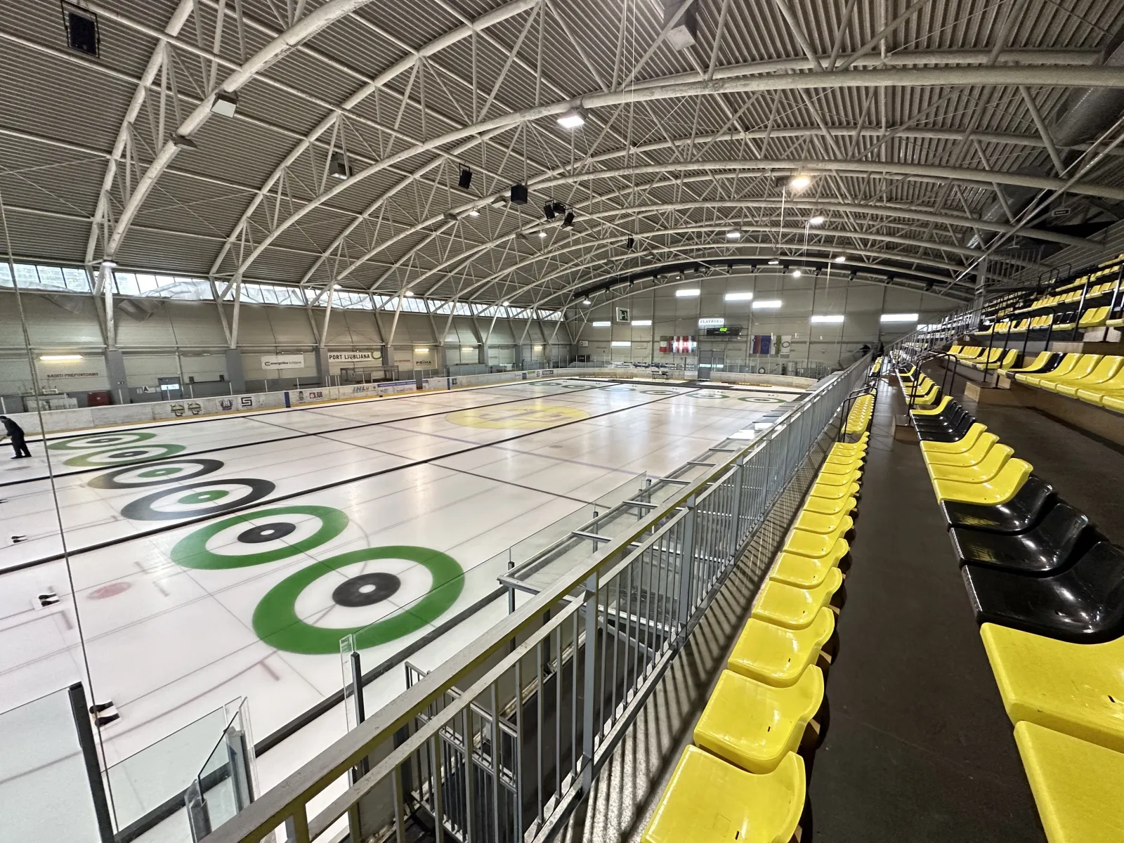 The Žalog ice hall seen from the stands, a curling target painted on the ice and the steel roof arching overhead. — Dvorana Zalog